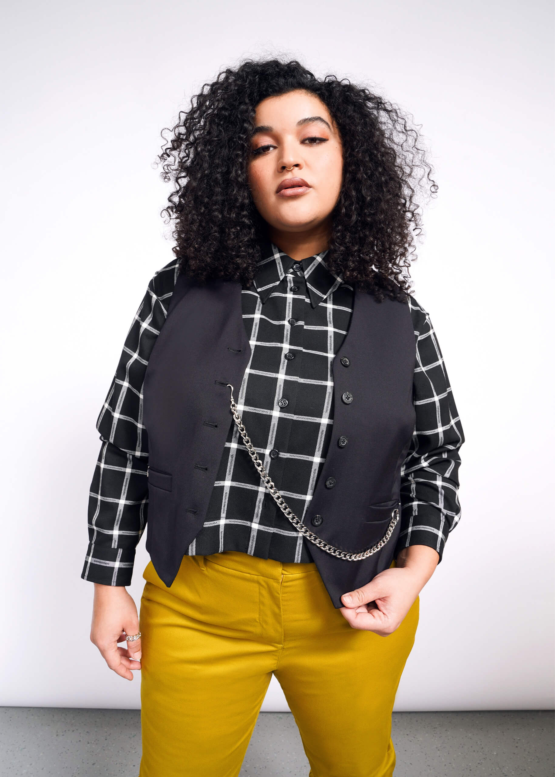 A curly-haired person models a Debbie Harry X Wildfang Vest over a black and white checkered shirt with mustard yellow trousers, set against a plain backdrop.