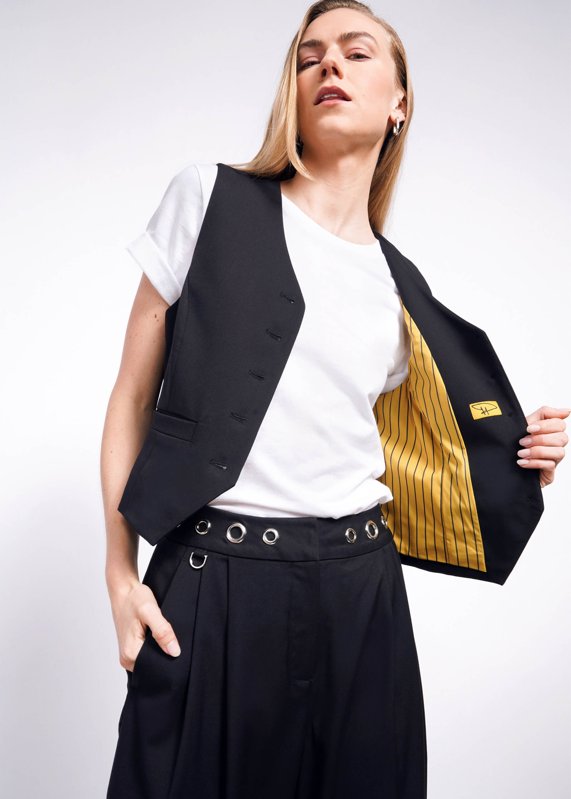 A person confidently poses in a white t-shirt, an open Debbie Harry X Wildfang Vest with yellow striped lining, and black grommet-adorned trousers against a plain background.