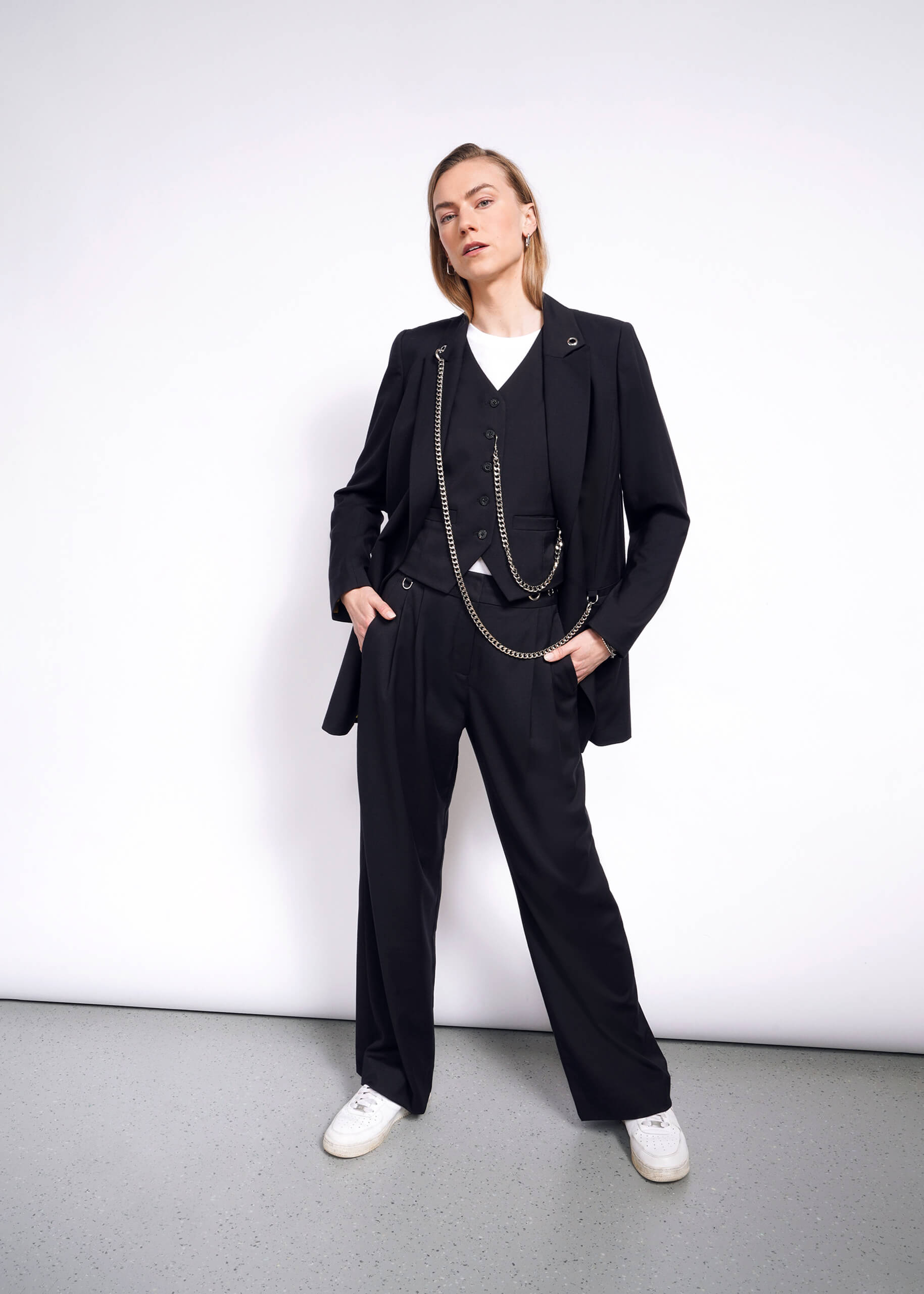 Posing against a white wall, the person wears a black suit with a Debbie Harry X Wildfang vest, white shirt, and sneakers. Hands in pockets, they strike a confident pose with a slight head tilt on the neutral gray floor.