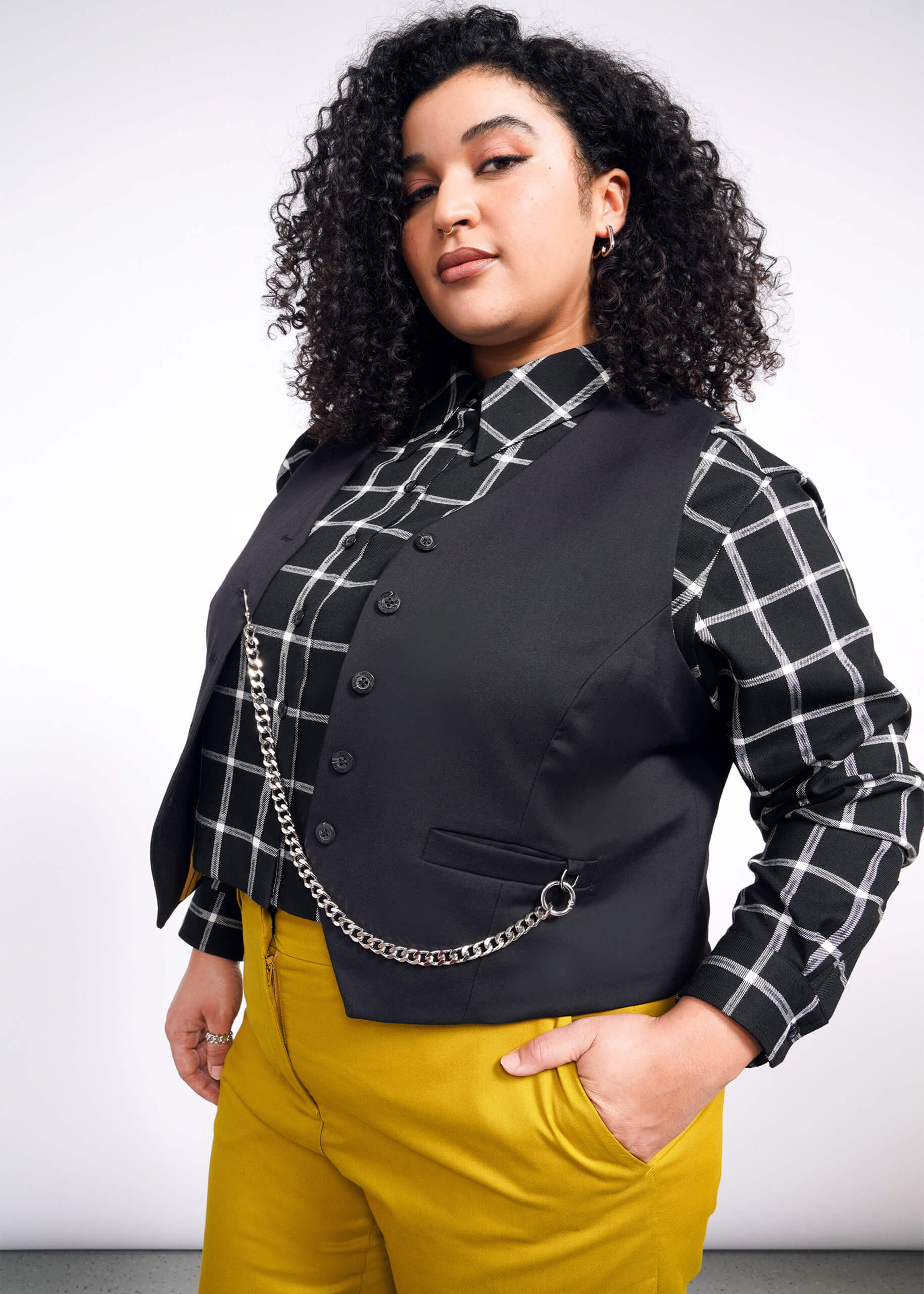 A person with curly hair, a black and white checkered shirt, a Debbie Harry X Wildfang Vest, and mustard yellow pants stands confidently against a simple background.