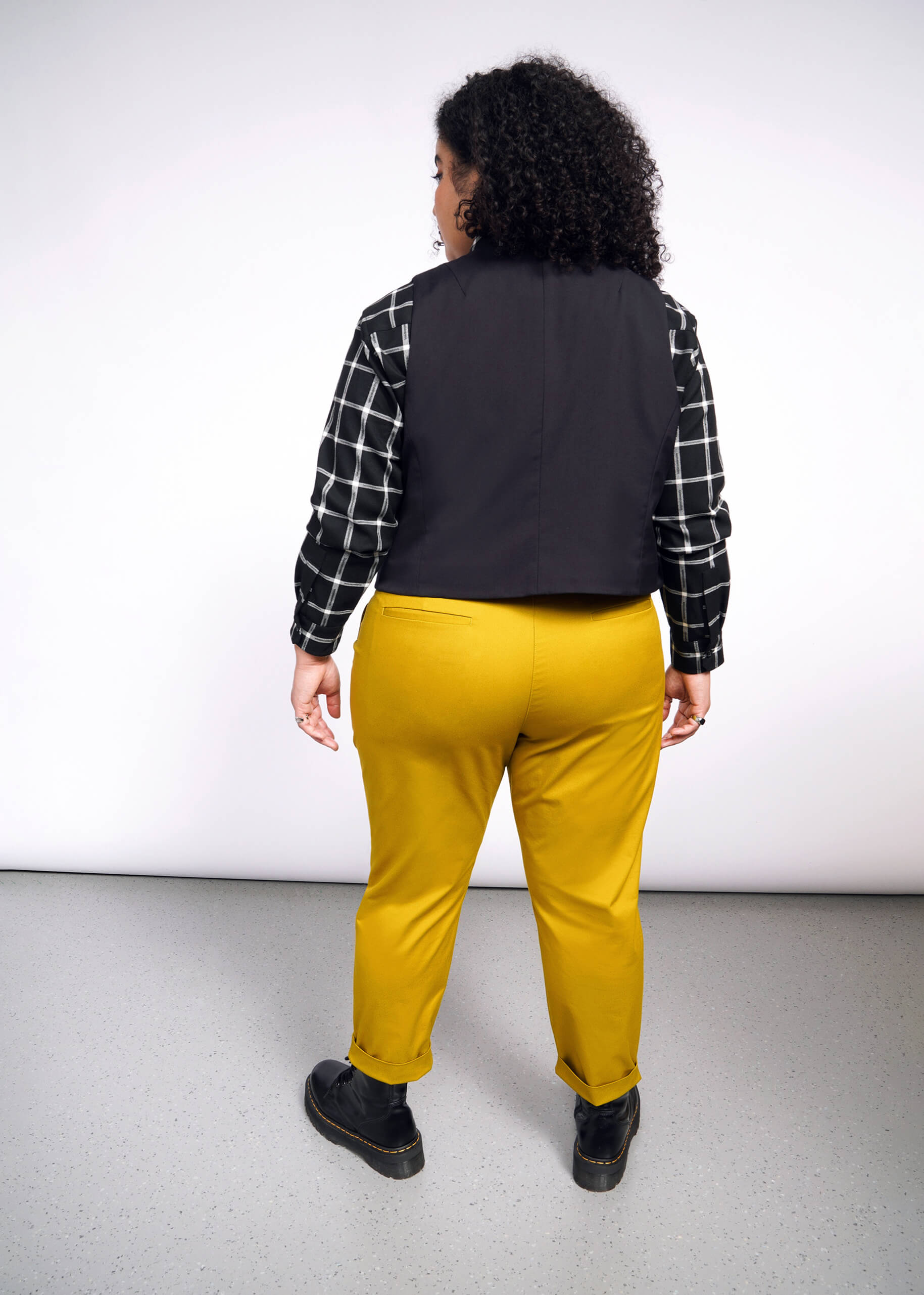 Rear view of a person with curly hair in a checkered shirt, Debbie Harry X Wildfang Vest, mustard yellow pants, and black shoes against a plain light background.