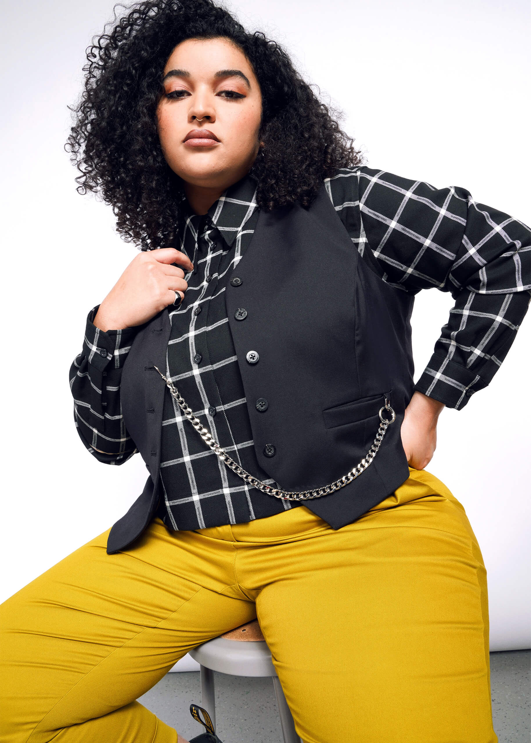 The person with curly hair sits confidently on a stool against a white backdrop, wearing a black and white checkered shirt, bright yellow pants, and the Debbie Harry X Wildfang Vest enhanced with a chain.