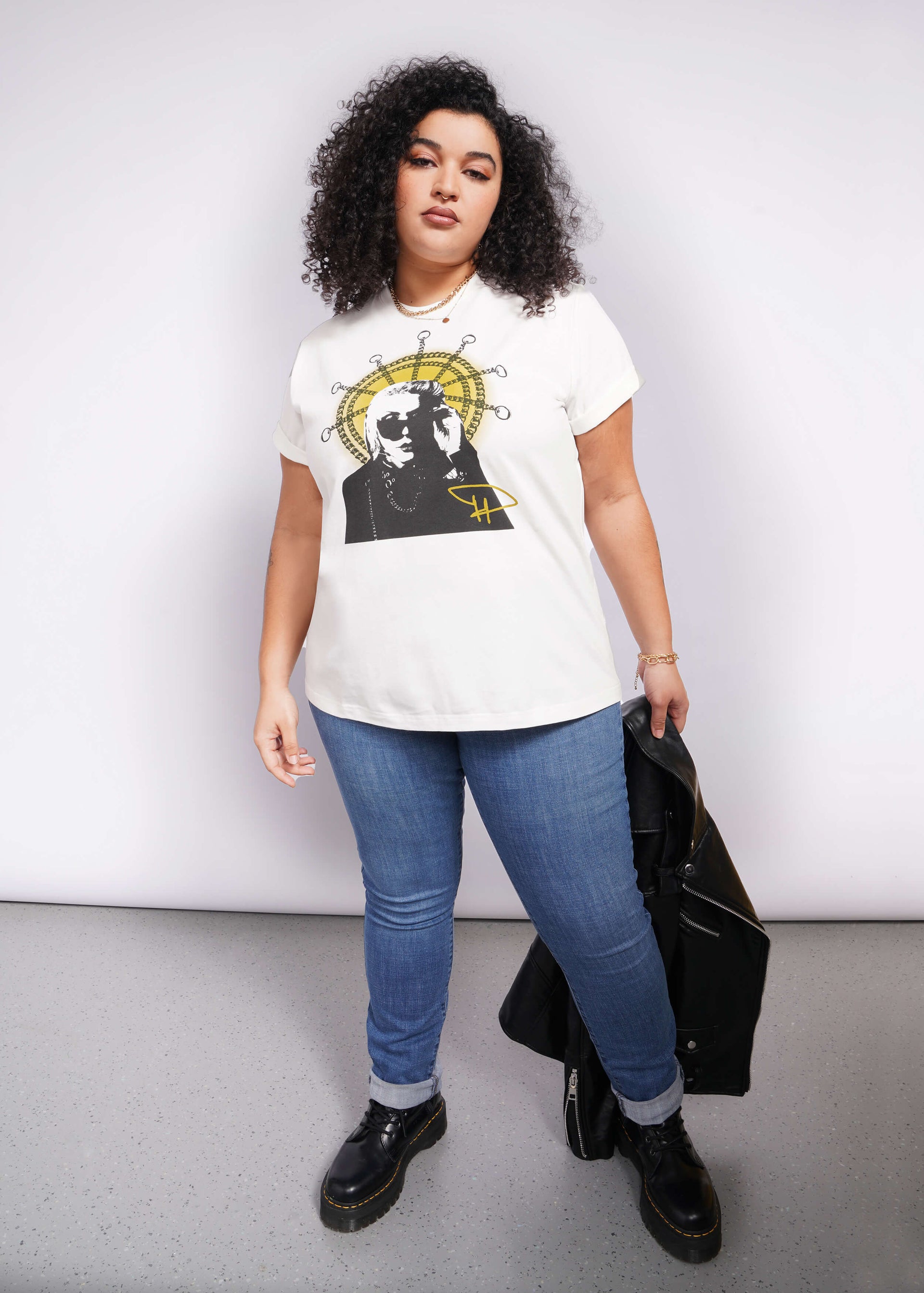 A person with curly hair confidently rocks a Debbie Harry X Wildfang Graphic Tee. Paired with blue jeans and black boots, they hold a black jacket against a plain backdrop, exuding an effortlessly cool vibe reminiscent of classic punk fashion.