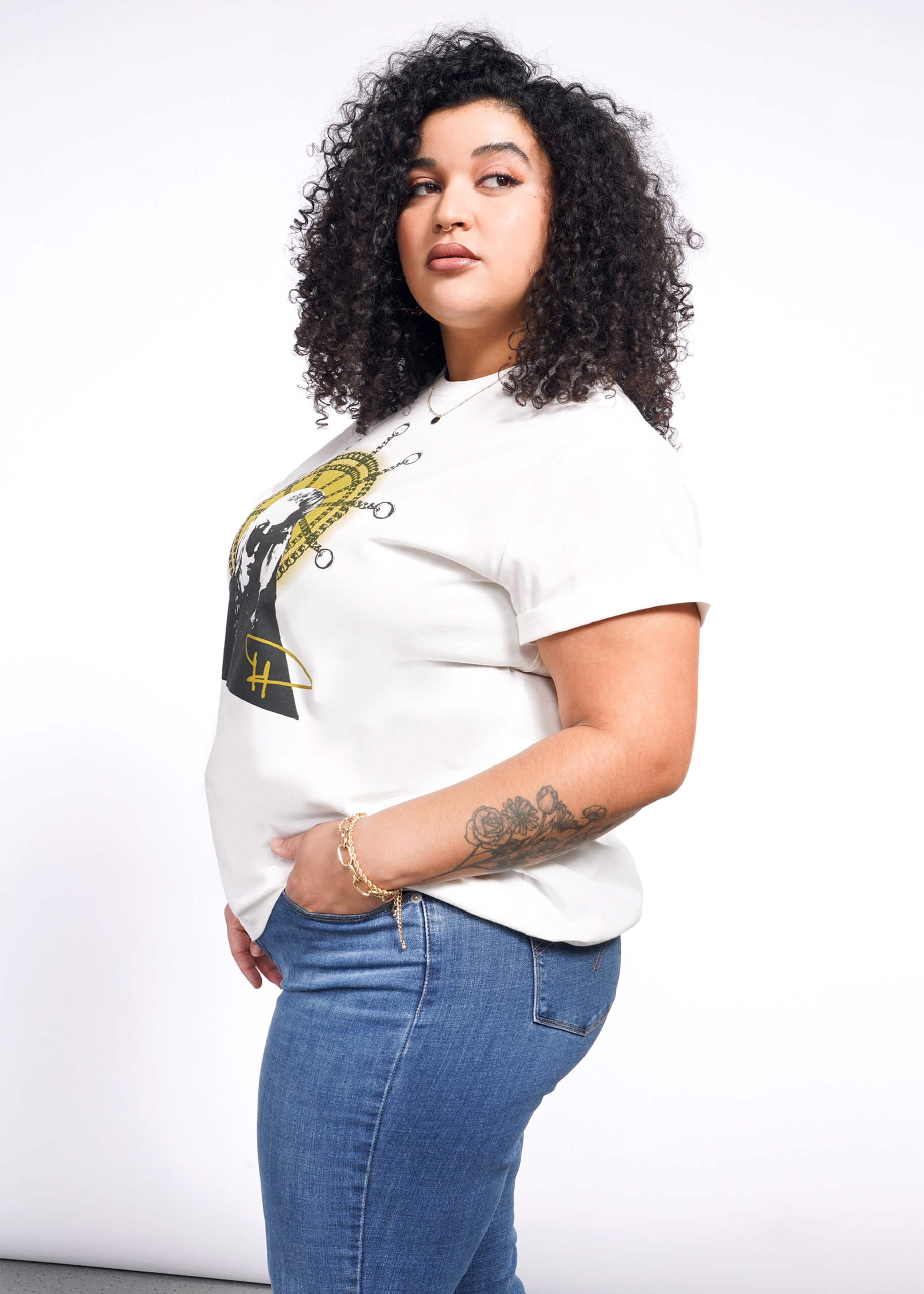 A person with curly hair stands sideways, donning a Debbie Harry X Wildfang Graphic Tee and blue jeans. Their hand is in their pocket, showcasing an arm tattoo. A plain white background accentuates their punk fashion style.