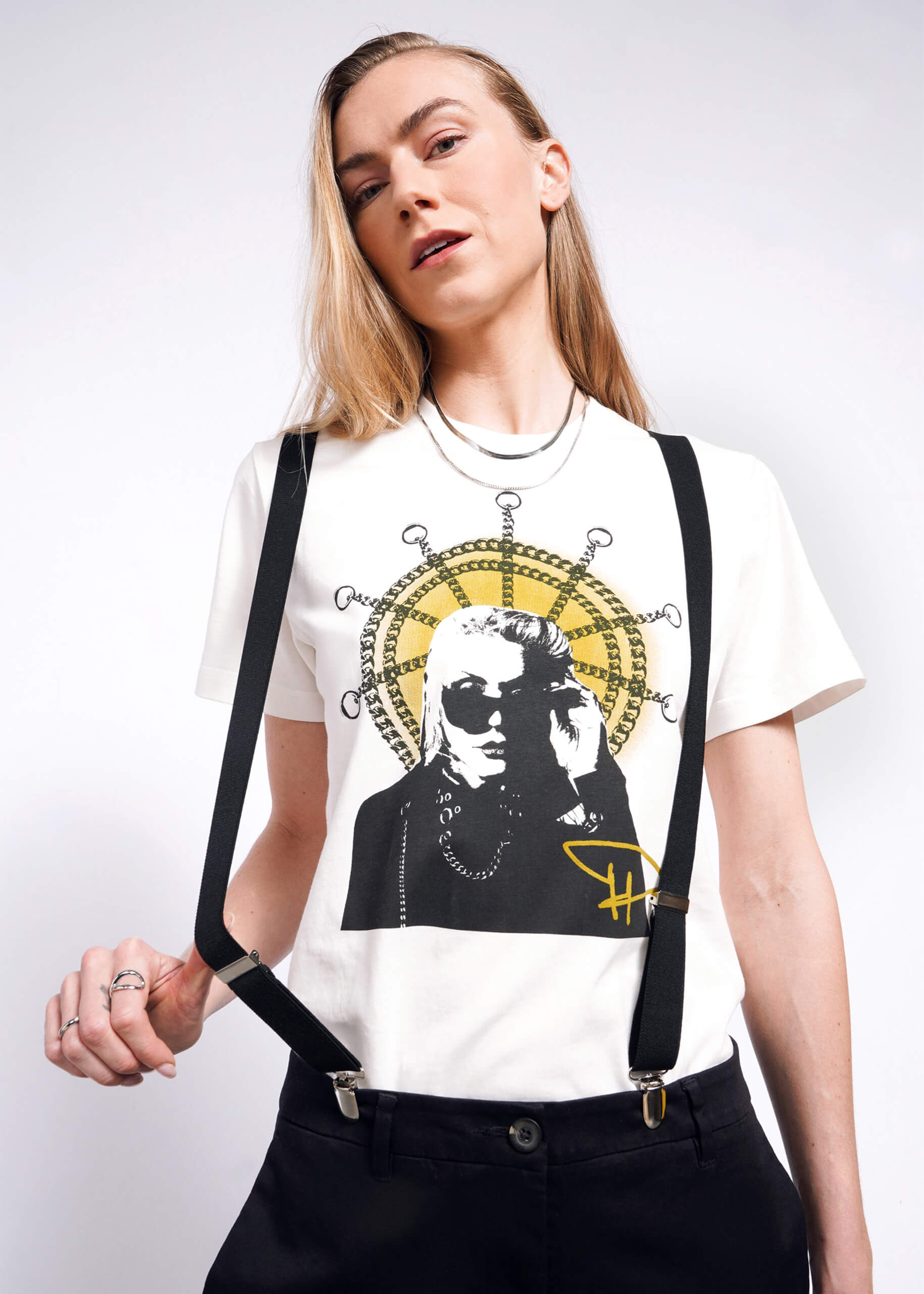 A person with long hair wearing the Debbie Harry X Wildfang Graphic Tee, featuring a graphic of someone in sunglasses, showcases punk fashion with layered necklaces and black straps against a plain background.