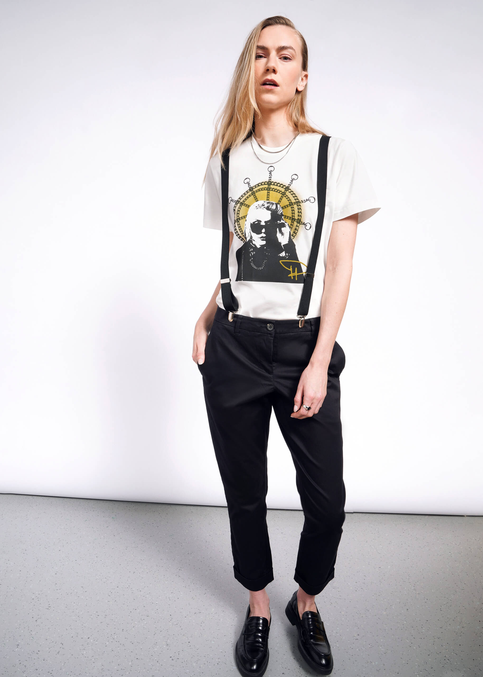 A person with long blond hair stands against a white background, wearing a Debbie Harry X Wildfang Graphic Tee. Black suspenders, pants, and loafers complete the punk look as they maintain a neutral expression with hands in their pockets.