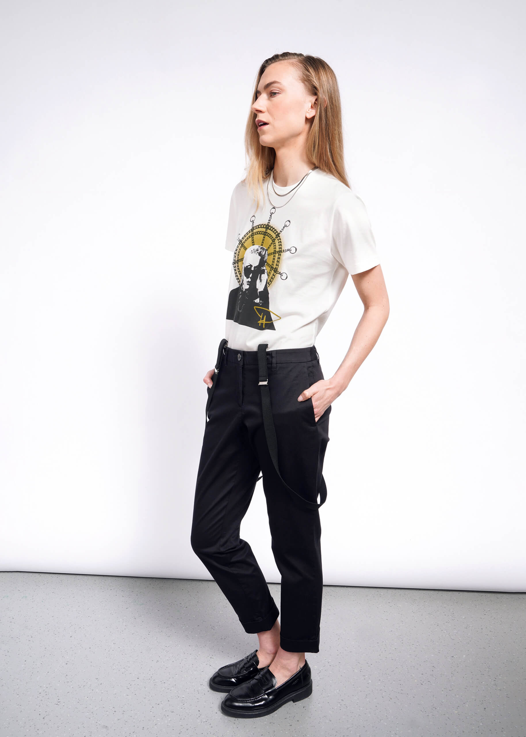 A person with long hair dons the Debbie Harry X Wildfang Graphic Tee, paired with black pants and suspenders. Against a light gray wall, hands in pockets, their shiny black shoes shine under the light, capturing punk fashion effortlessly.