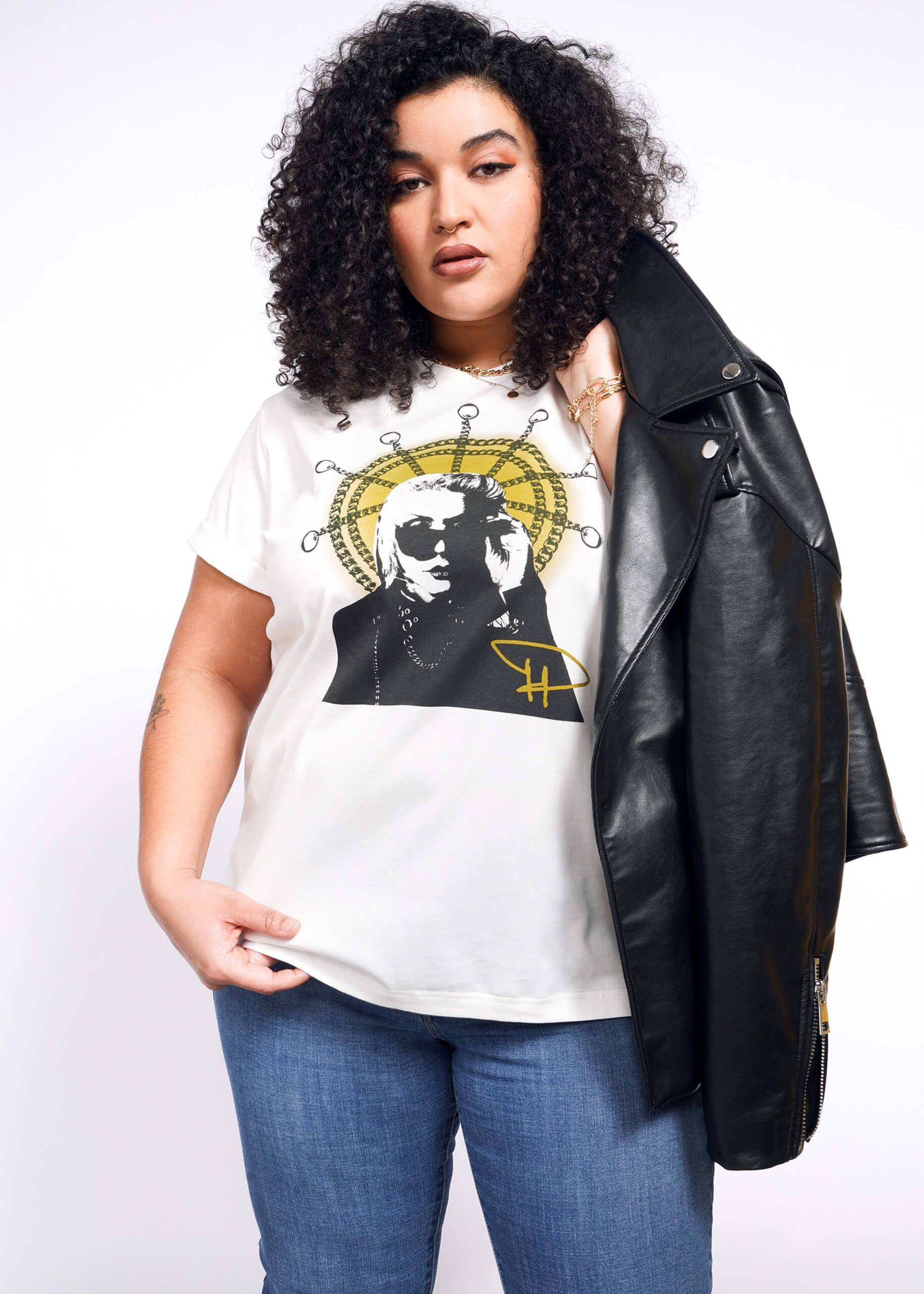 With curly hair, a person sports a Debbie Harry X Wildfang Graphic Tee featuring an abstract design while slinging a black leather jacket over one shoulder, exuding punk fashion against a plain white backdrop.