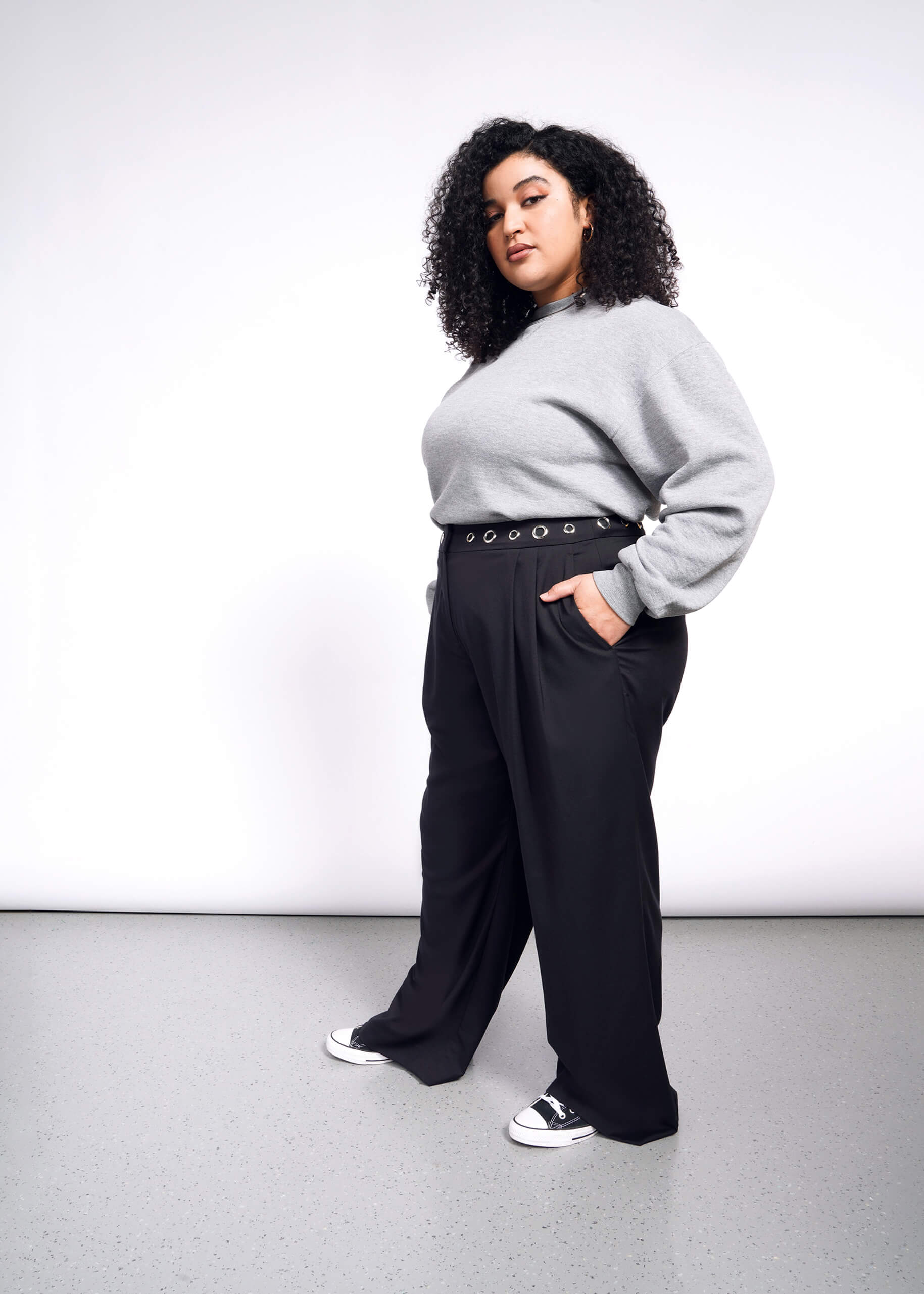 Curly-haired and confident, a person poses against a white background in Debbie Harry X Wildfang Wide Leg Trousers, paired with a grey sweater and black sneakers. With hands in pockets, they look at the camera with a relaxed posture.