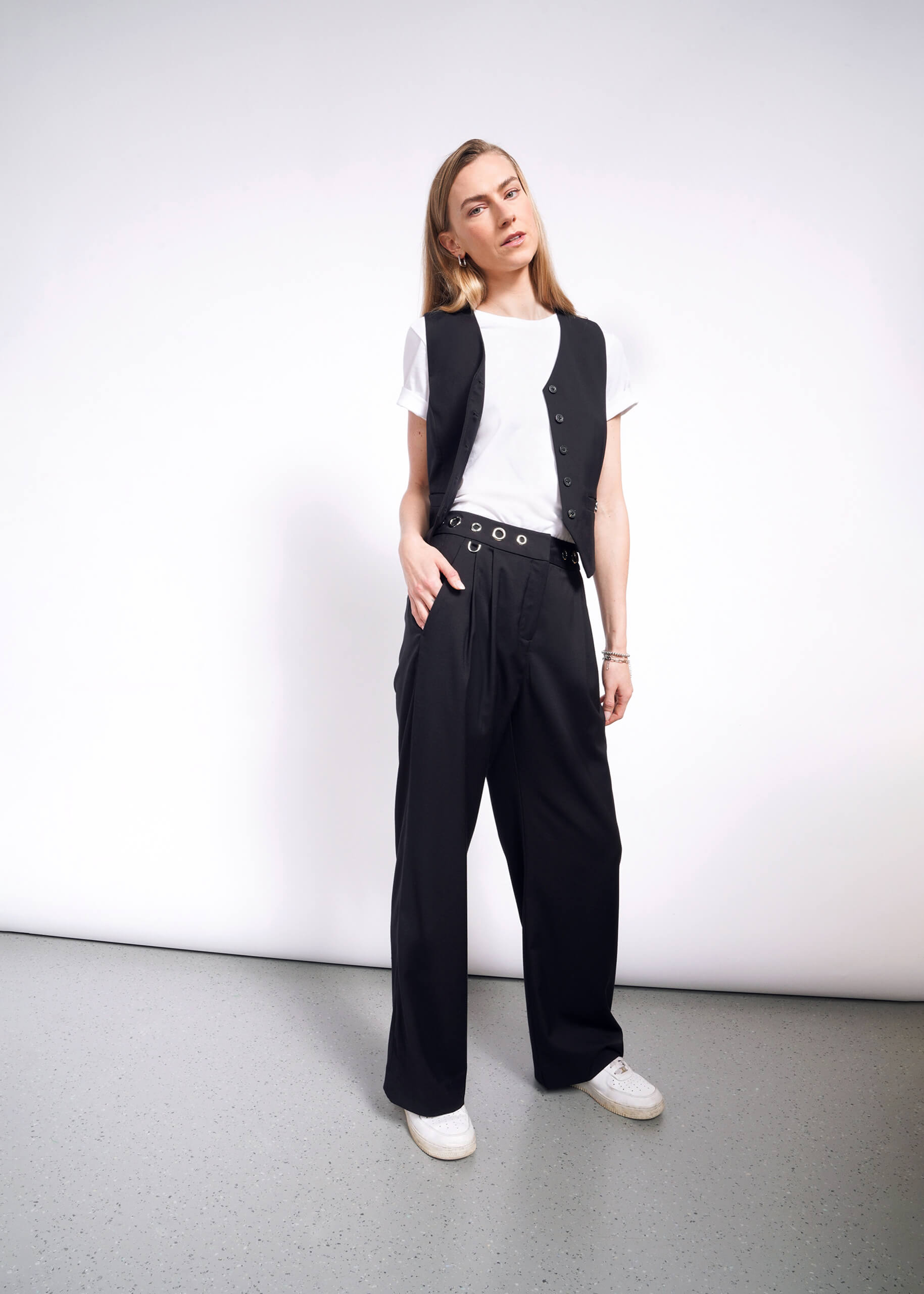 A person in a white t-shirt, black vest, and Debbie Harry X Wildfang Wide Leg Trouser with a grommet belt stands relaxed in a minimalist space with a neutral background, wearing white sneakers from the Debbie Harry collection.