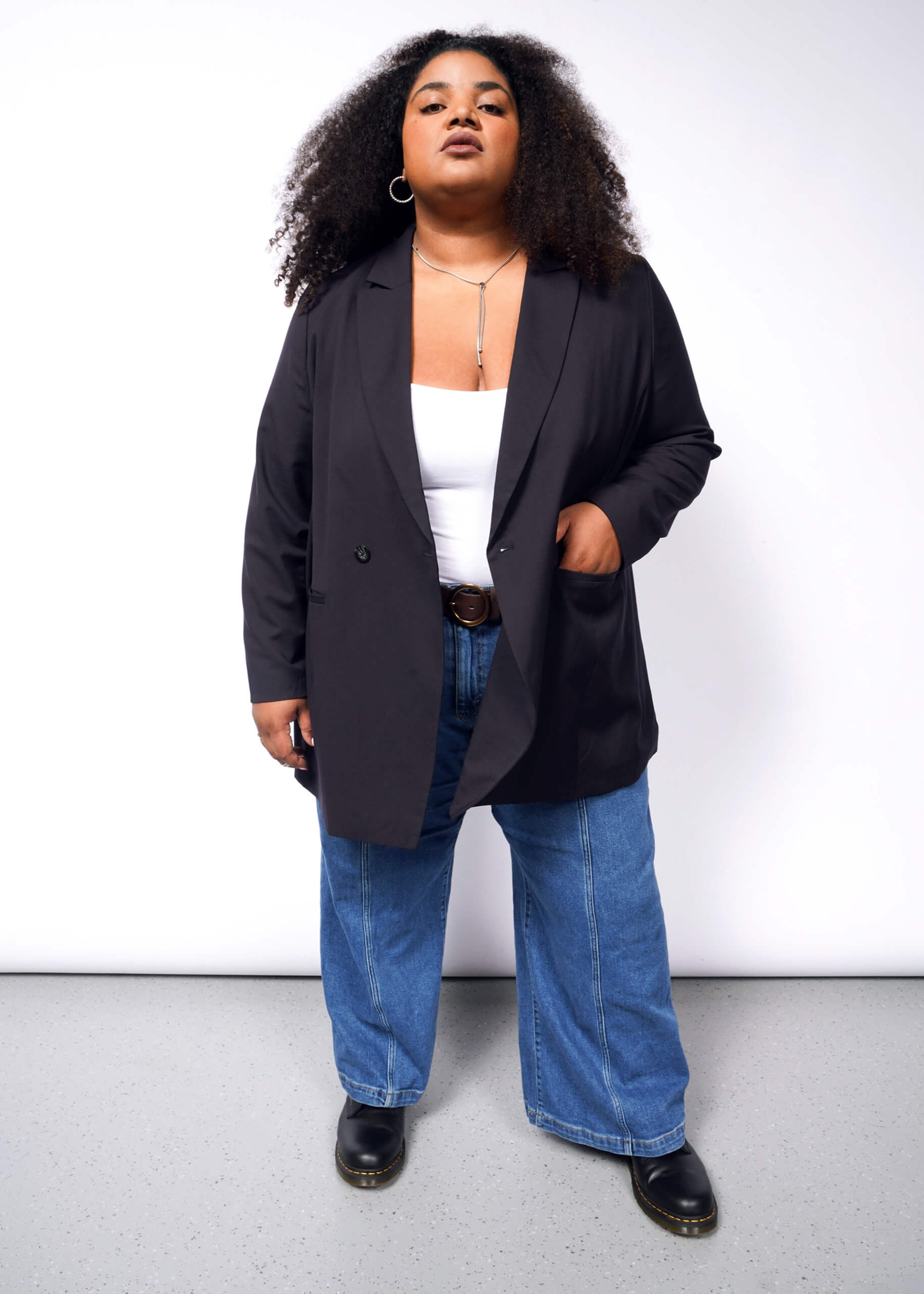 A person with curly hair is wearing The Empower Double Breasted Blazer in black over a white top, paired with blue jeans and black shoes. Their left hand rests in a pocket as they stand against a plain white background.