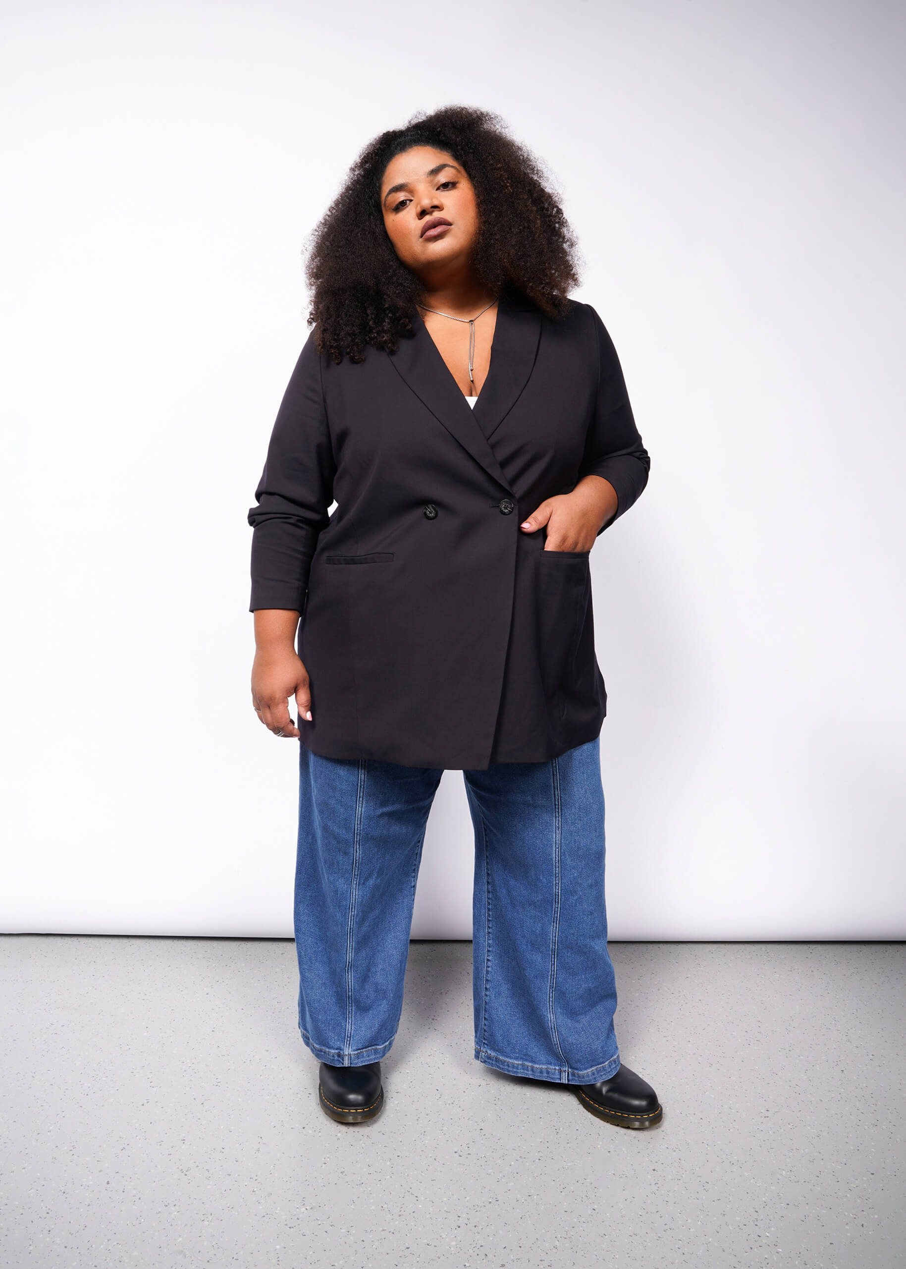 Against a plain backdrop, a person with curly hair confidently poses, wearing The Empower Double Breasted Blazer in black, paired with blue wide-leg jeans and black shoes. With one hand in their pocket, they look directly at the camera.