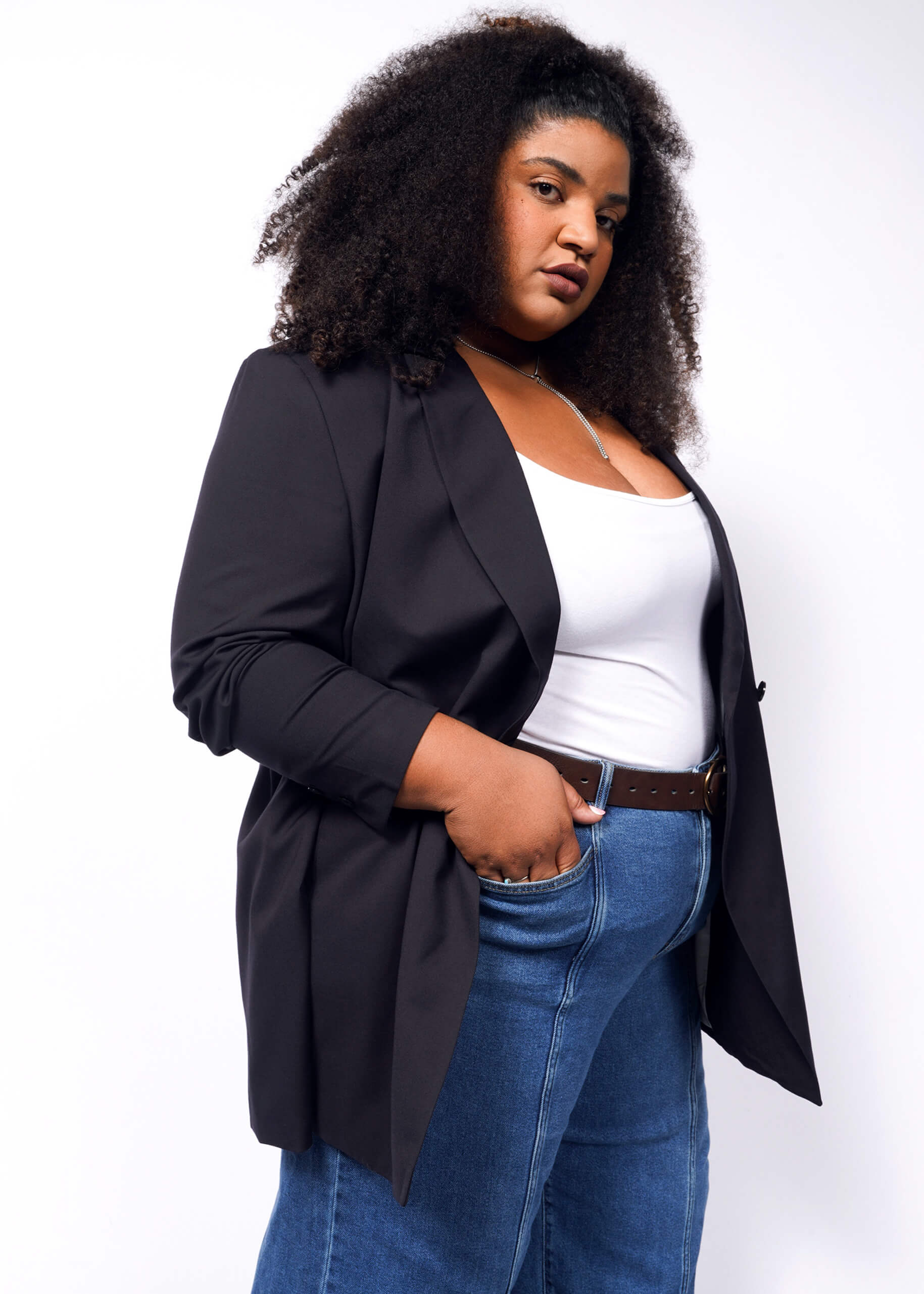 A person with long curly hair stands confidently before a white backdrop, wearing The Empower Double Breasted Blazer in black over a white top, paired with blue jeans and a brown belt. One hand rests in their pocket, exuding poise and style.