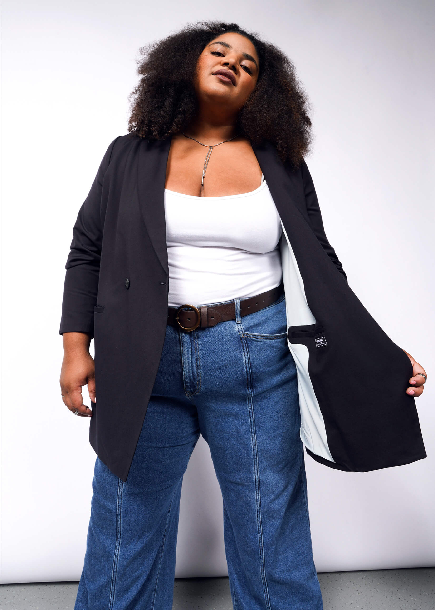 A person with curly hair confidently stands against a plain background wearing The Empower Double Breasted Blazer in black over a white top and blue jeans, showcasing the tailored fit and elegant inner lining of the open blazer.