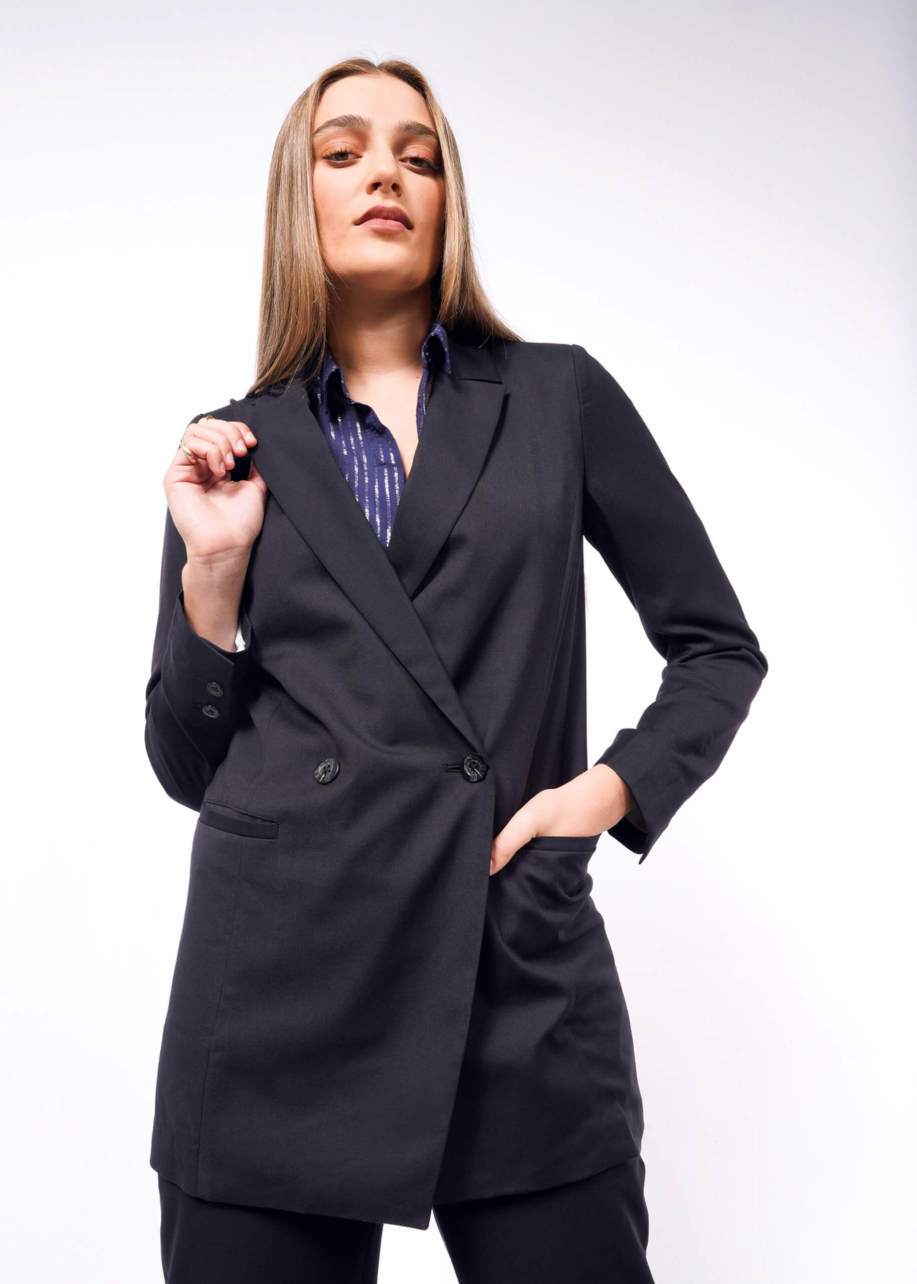 Wearing The Empower Double Breasted Blazer over a striped shirt, a person stands confidently against a plain white background. With one hand on their hip and the other adjusting the blazers lapel, they embody modern sophistication.