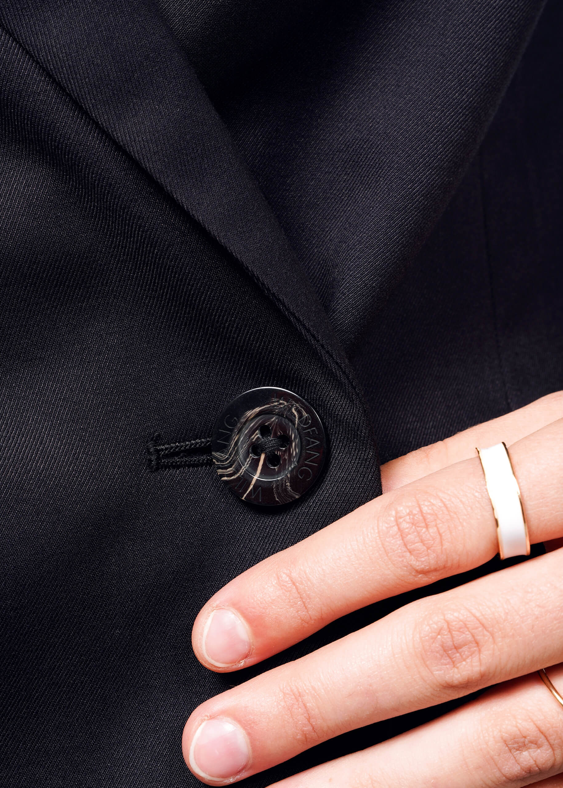 A hand touches The Empower Double Breasted Blazer. Focus is on a round logo-engraved button. A simple wide ring on the index finger complements the blazers elegant long-line design.