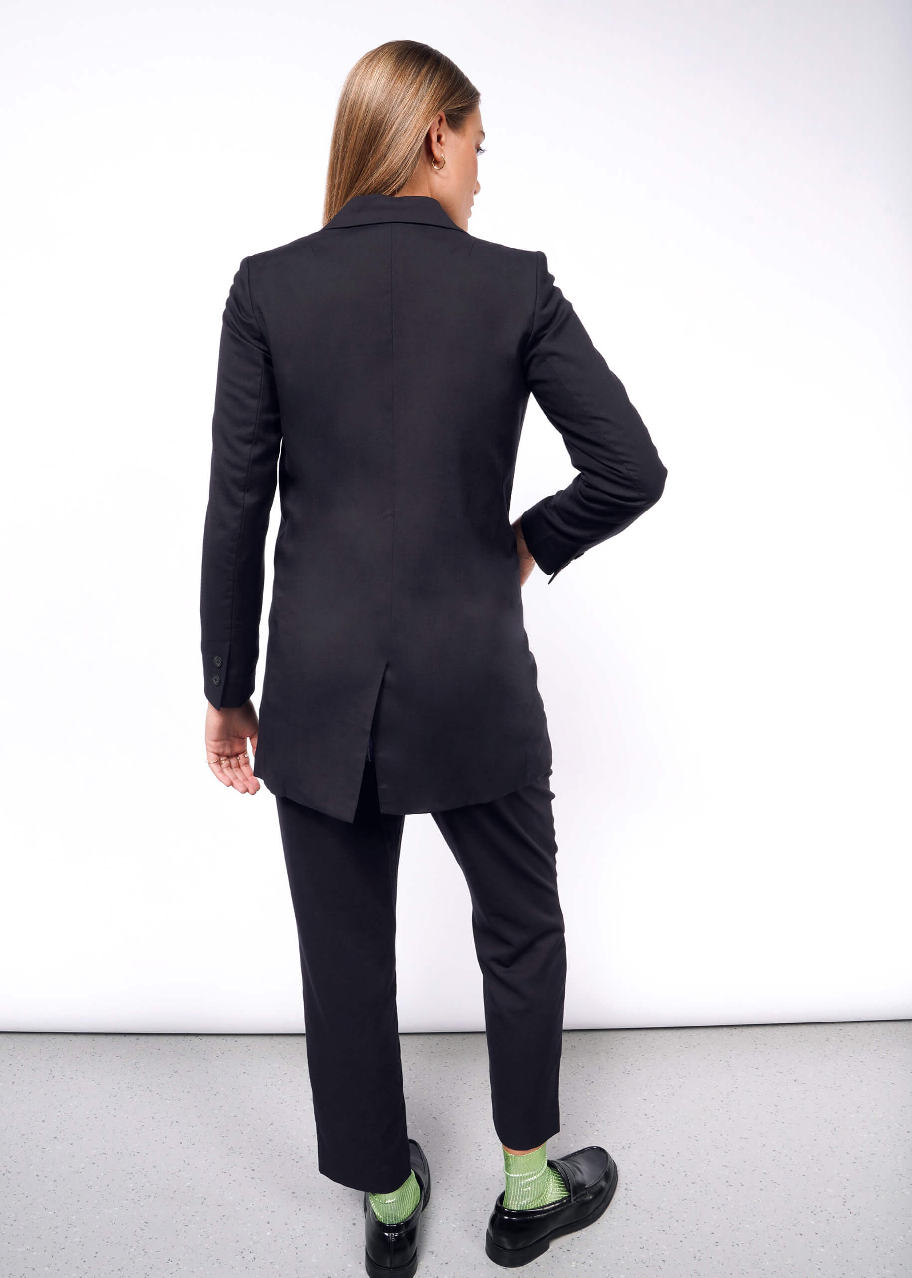 A person with long hair stands facing away, showcasing The Empower Double Breasted Blazer paired with green socks. The tailored fit creates a striking silhouette in the brightly lit room with its gray floor and white background.