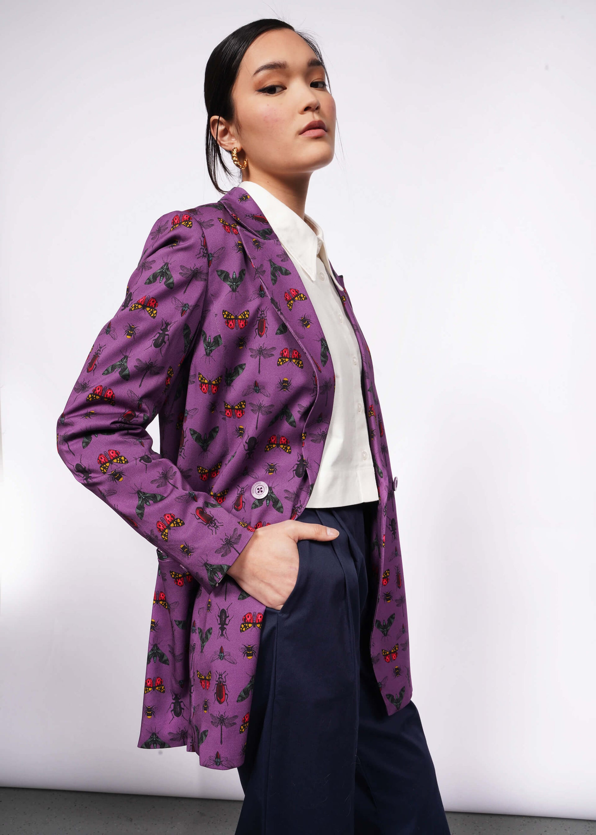 Side view of person with black hair and hand in trouser pocket wearing purple bug printed blazer unbuttoned with white button up shirt underneath