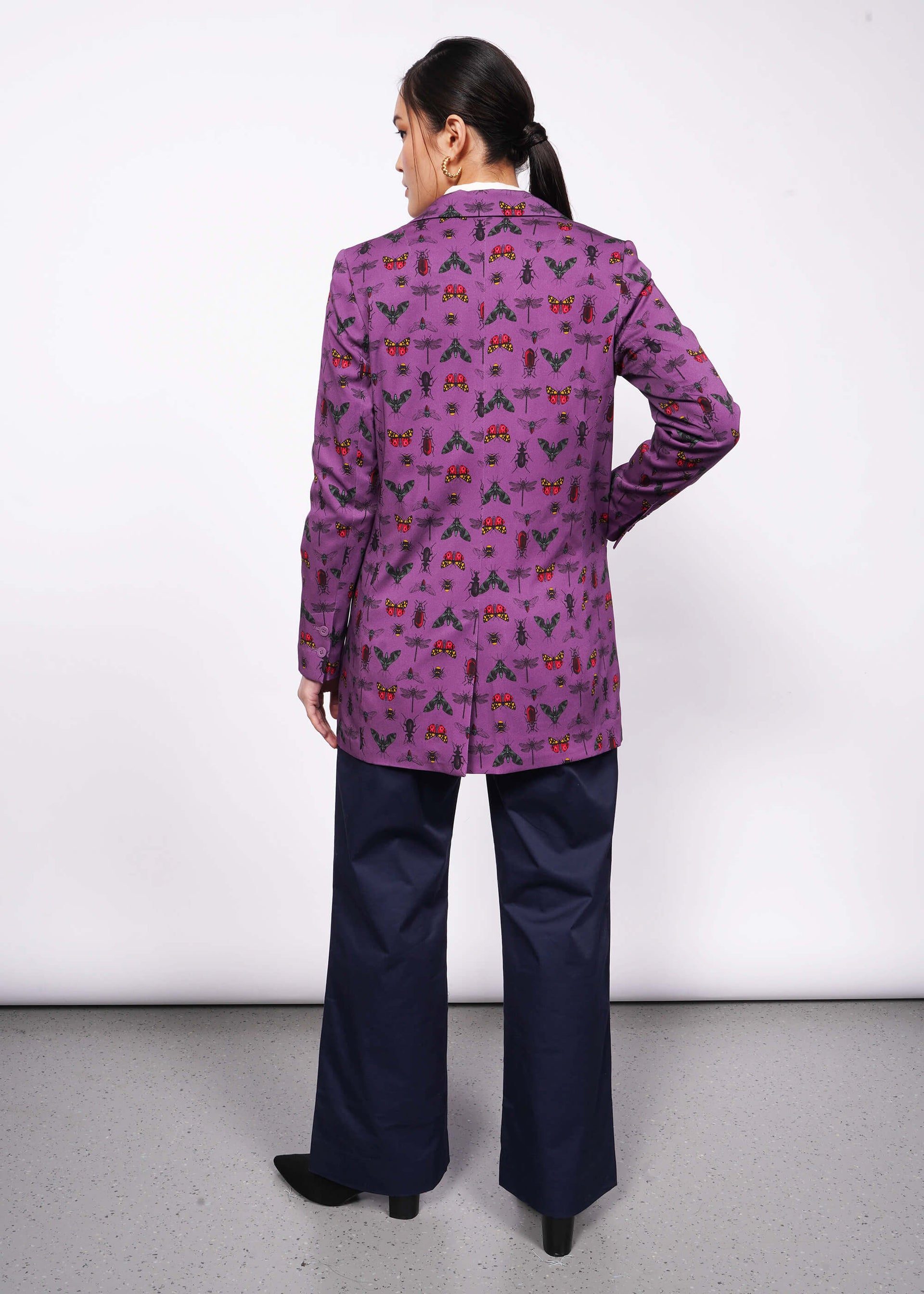 Back view of person with black hair and hand in trouser pocket wearing purple bug printed blazer with navy trouser and black heeled boots.