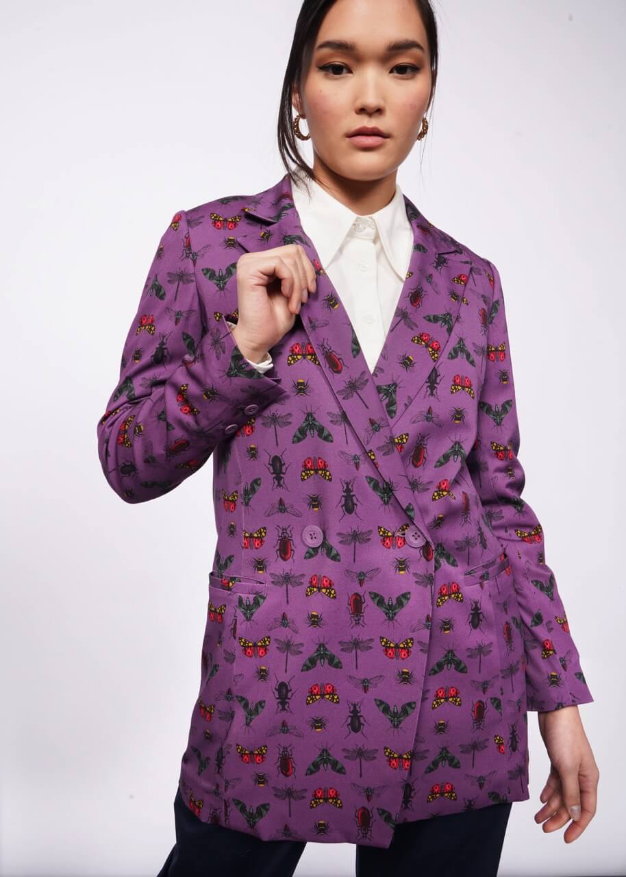 Person with black hair looks at camera holding the lapel of their purple blazer with a bug print on it. They are wearing a white shirt underneath. Neutral background.