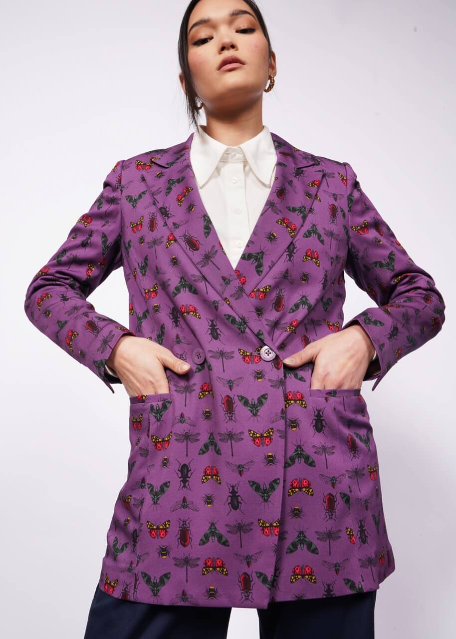 Front view of person with black hair and hands in blazer pockets wearing purple bug printed blazer with white button up shirt underneath