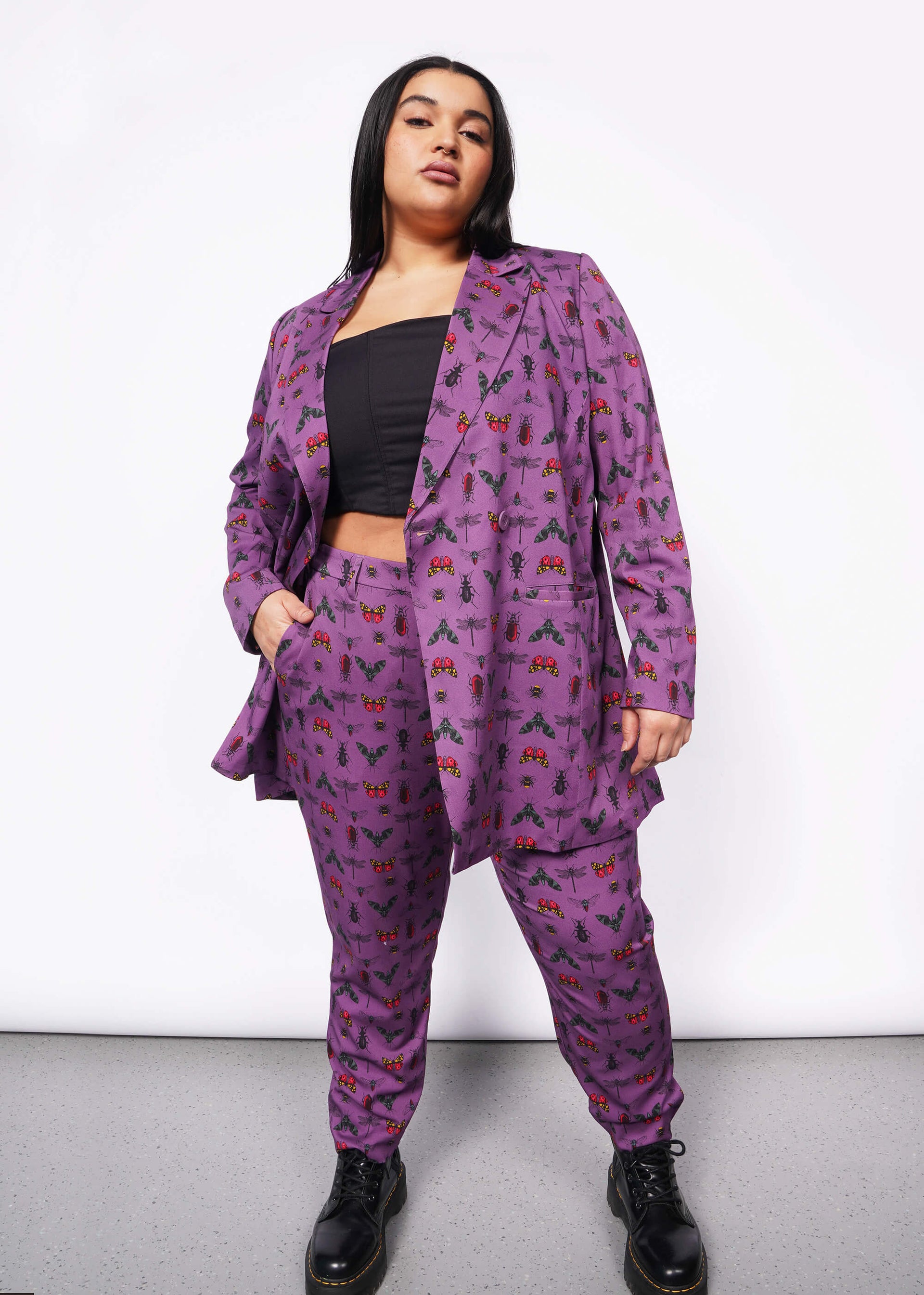 Front view of person with long black hair and nose ring looking at camera with hand in pant pocket. Blazer is unbuttoned and is purple with bug print. They are wearing black corset underneath blazer, a matching pant, and a black boot.