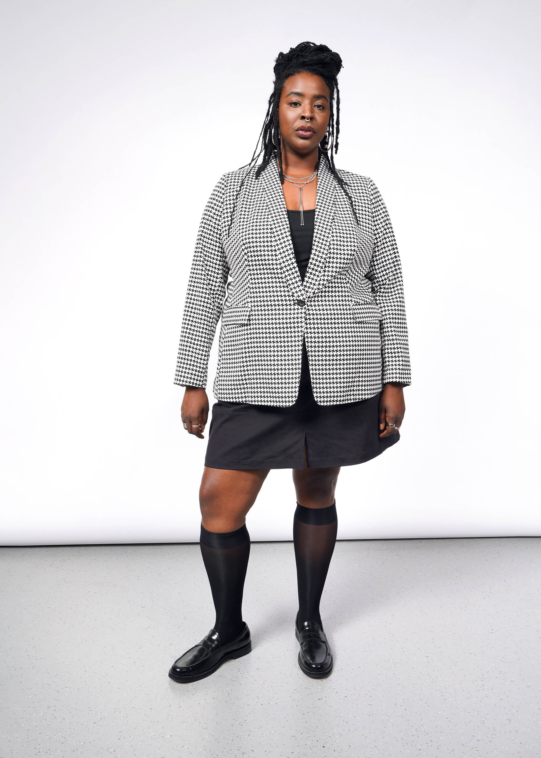 Wearing The Empower Houndstooth Tux Blazer in black and white with a tailored fit, a person confidently pairs it with a black skirt, knee-high socks, black loafers, and flaunts long braided hair while posing against a simple white wall and gray floor.