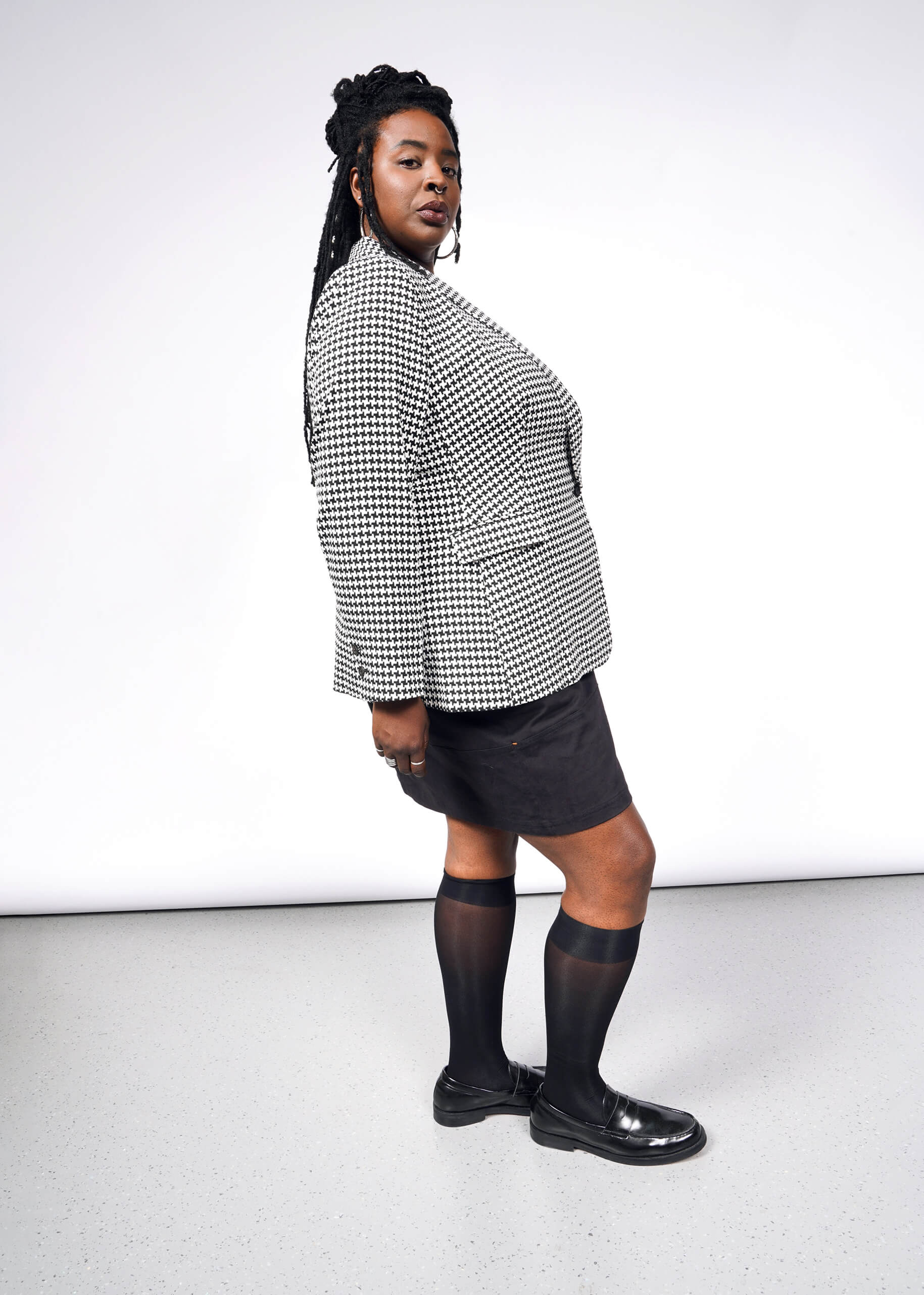 A person stands sideways, showcasing The Empower Houndstooth Tux Blazer with deep pockets. The tailored fit complements the black skirt and knee-high socks with shiny shoes, while their long braided hair cascades majestically in the well-lit studio against a plain backdrop.