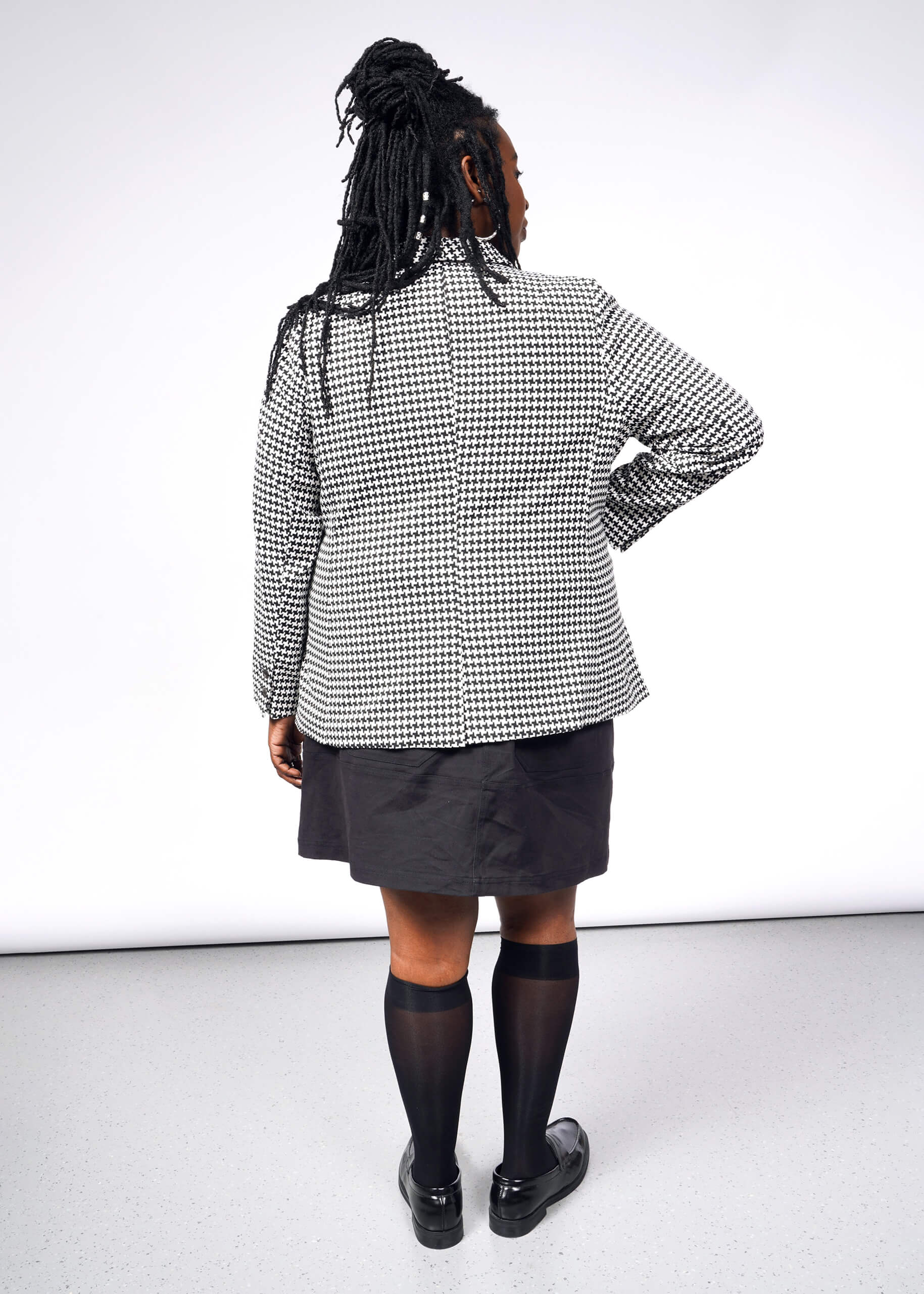 A person with long braided hair faces away, wearing The Empower Houndstooth Tux Blazer, a dark skirt, knee-high black socks, and black loafers. The tailored fit enhances their style against the plain white background.