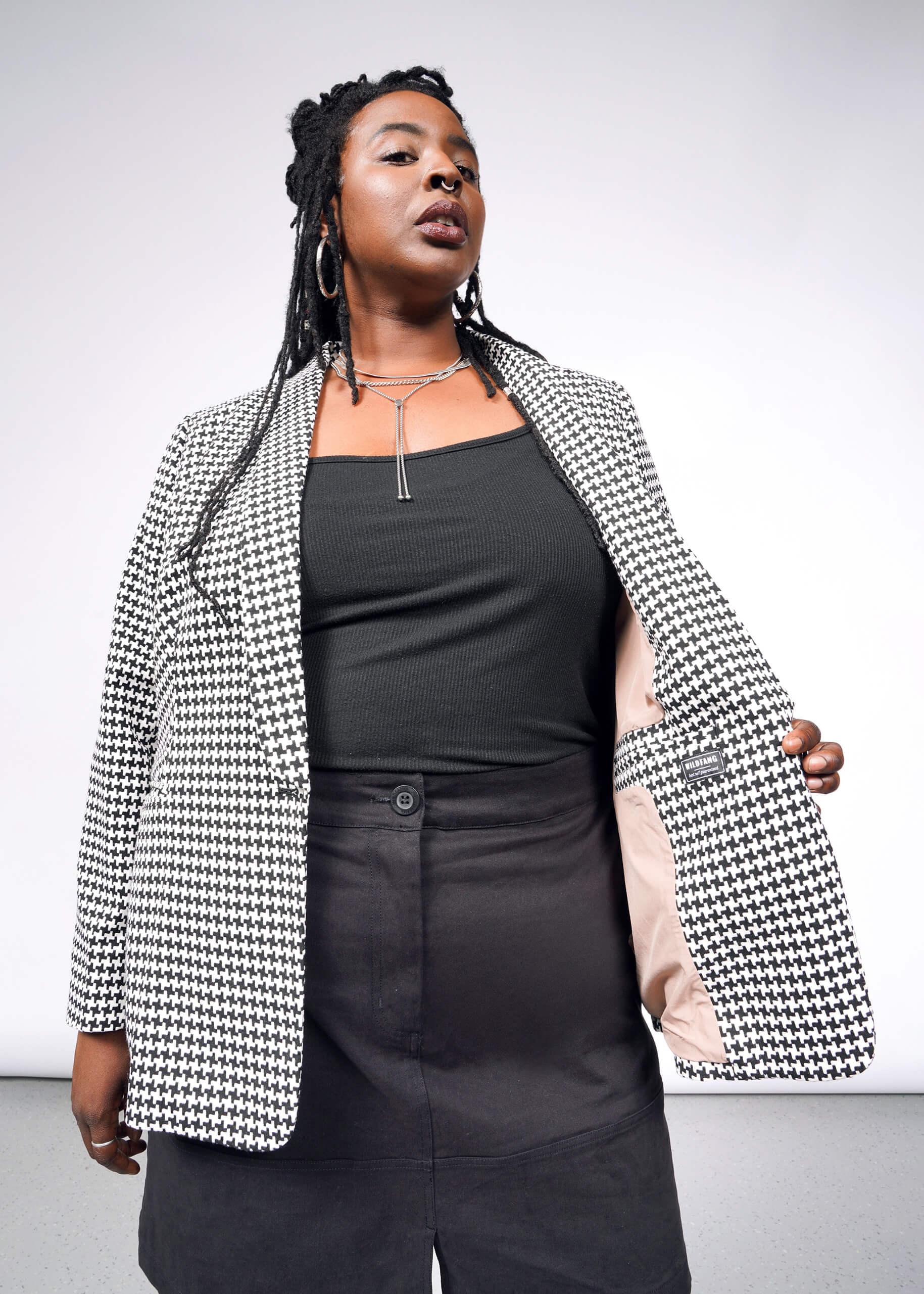 In The Empower Houndstooth Tux Blazer, a person exudes confidence with its tailored fit, worn over a black top and skirt. Their long braided hair, hoop earrings, and silver necklace accentuate the look. The open blazer reveals a beige lining against a plain background that highlights their style.