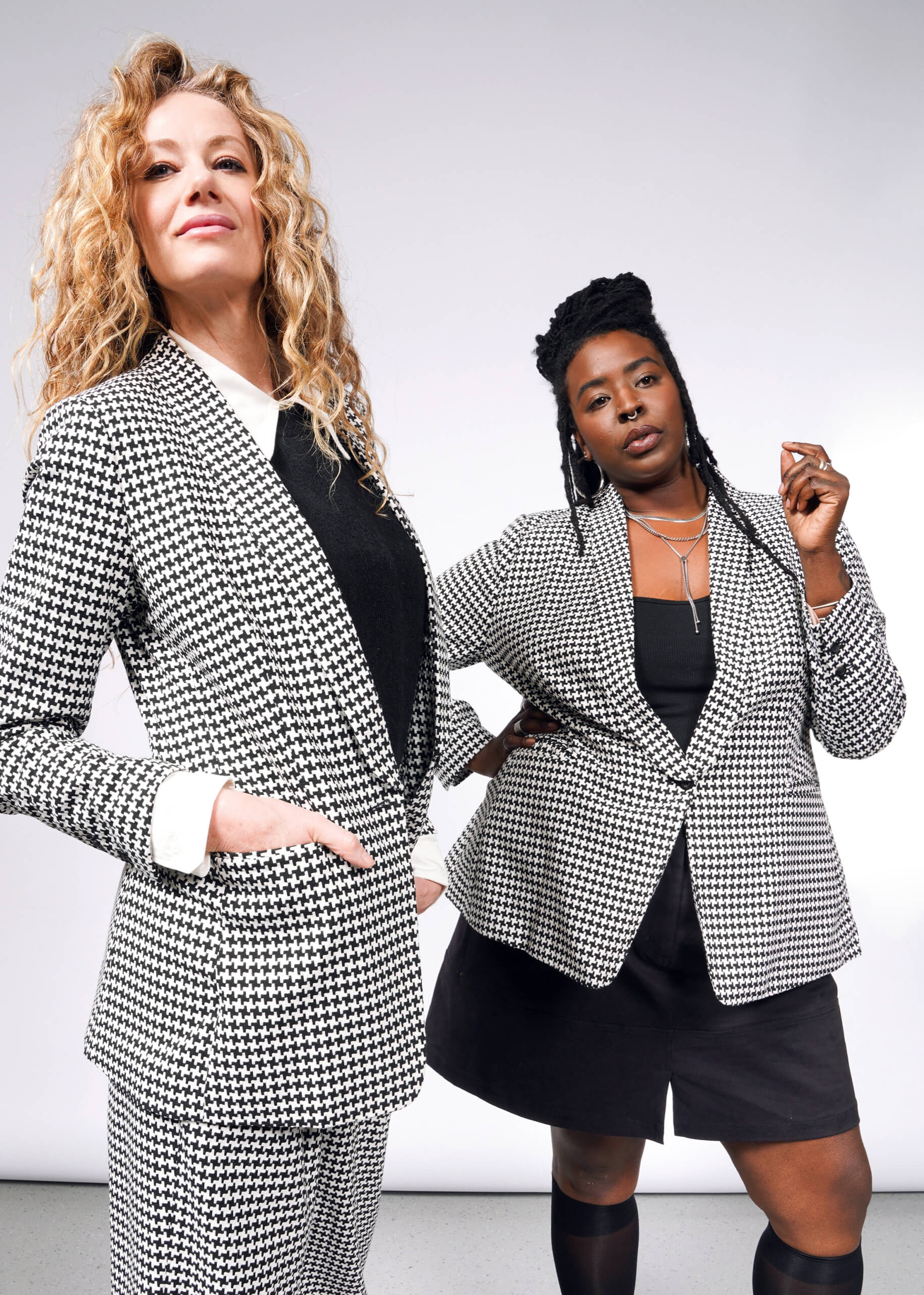 Two women in The Empower Houndstooth Tux Blazer pose confidently. The woman on the left sports curly blonde hair, and the woman on the right with braided hair pairs her look with a black top and skirt. Their assertive stance is enhanced by a stretch lining for added comfort.