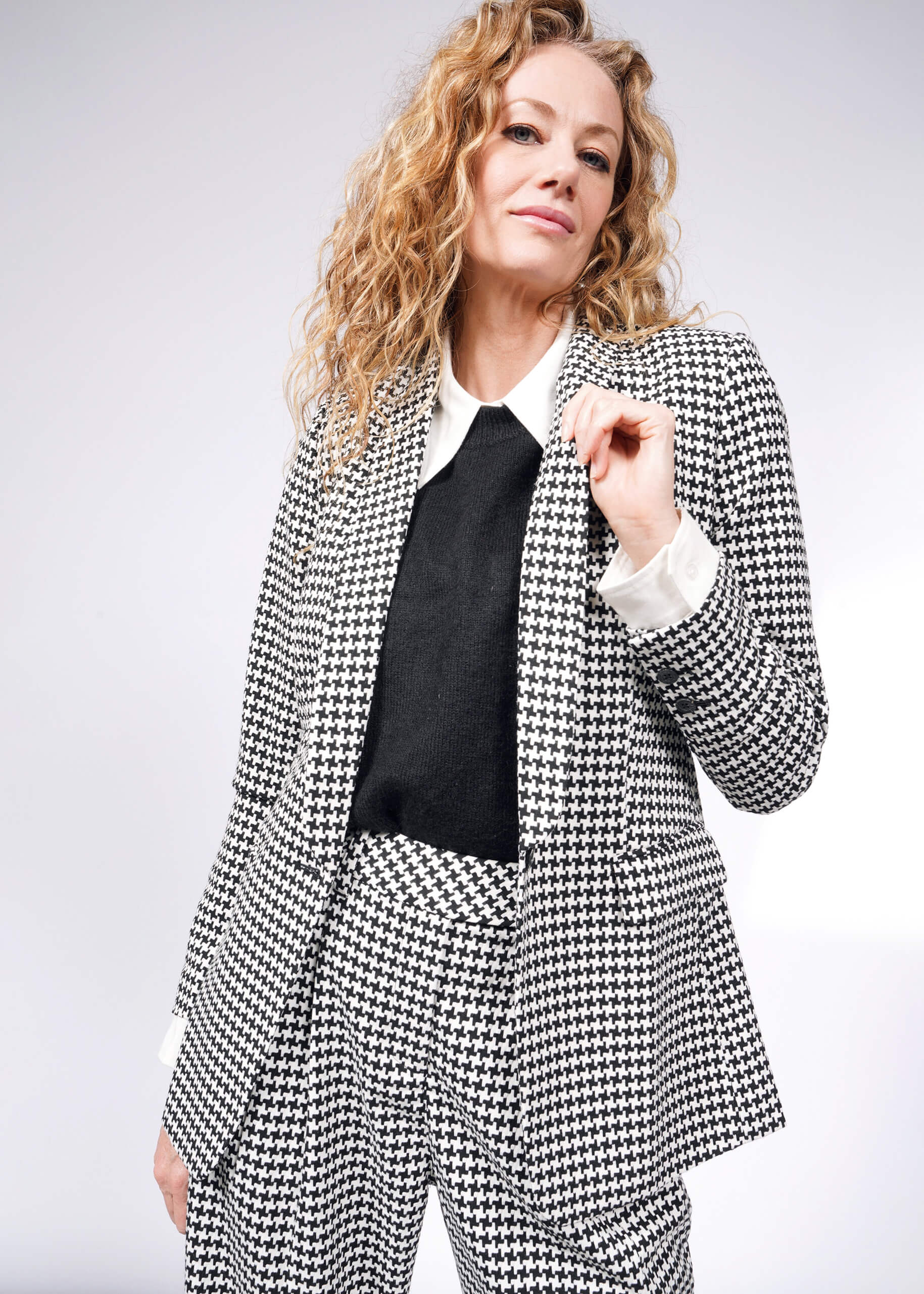 A woman with curly hair confidently poses in The Empower Houndstooth Tux Blazer, holding the lapel. Her relaxed expression is enhanced by the blazers stretch lining, while a light gray background complements her stylish look.