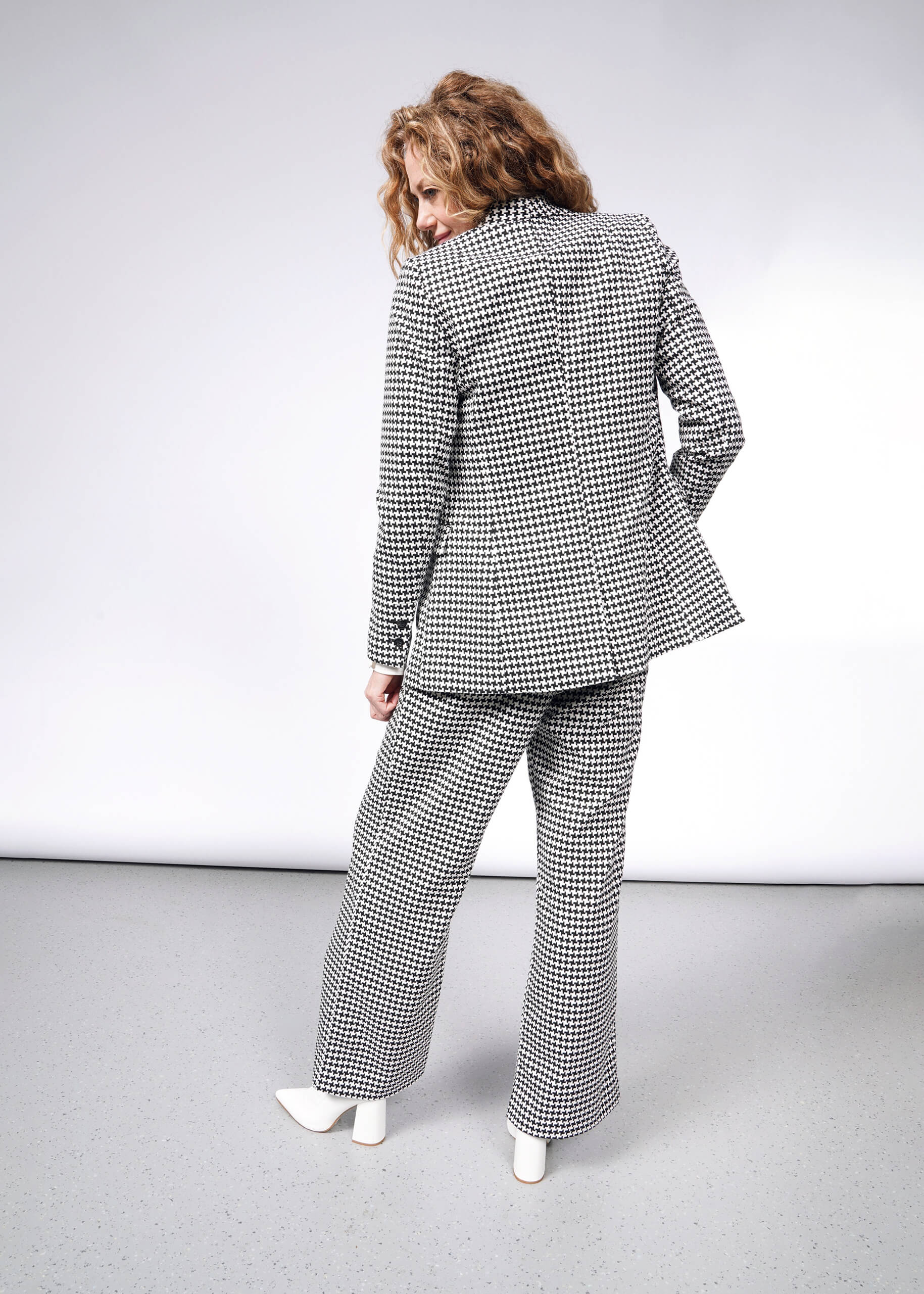 A person with curly hair stands with their back to the camera wearing The Empower Houndstooth Tux Blazer and white shoes; the background is a simple gray gradient.