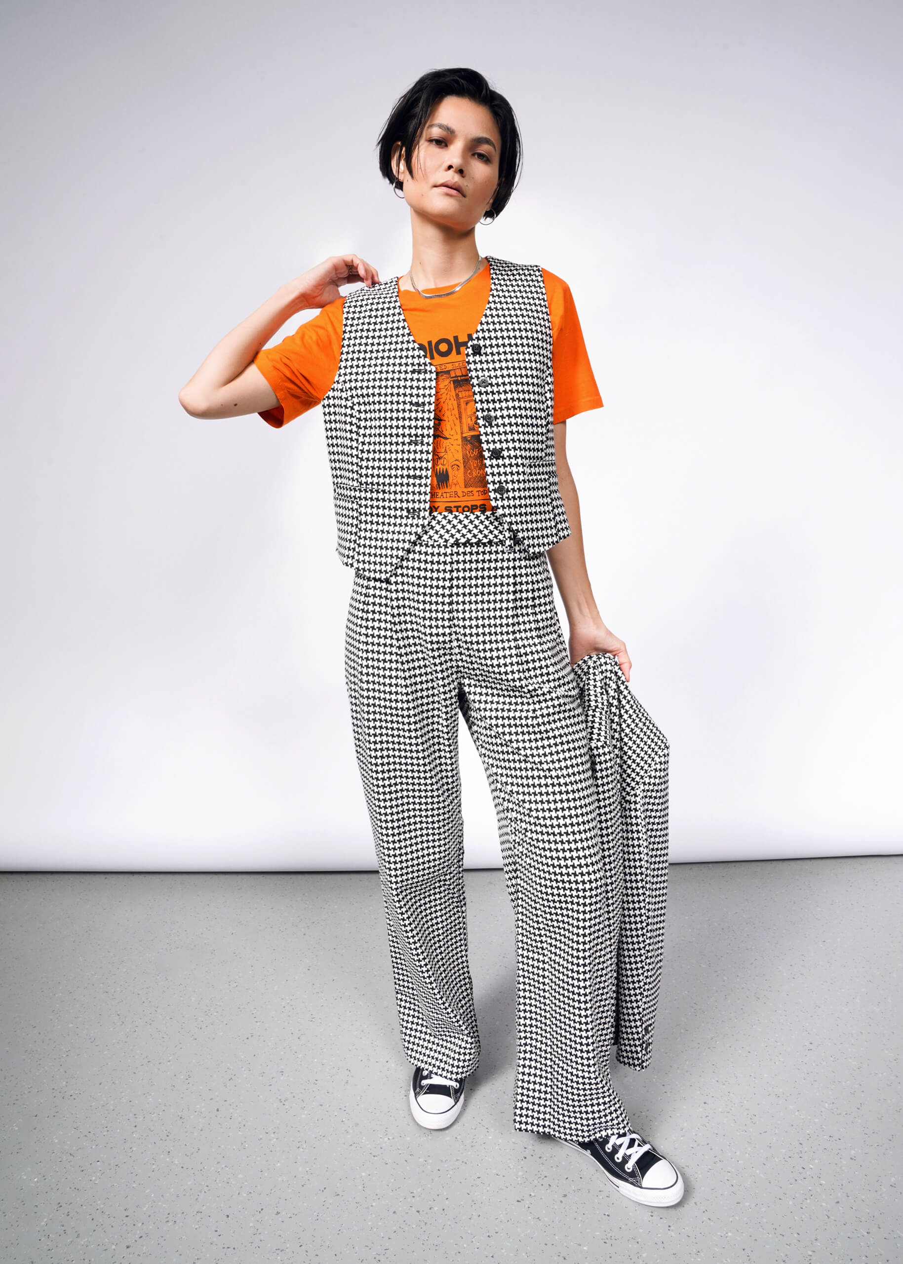 A person stands confidently against a plain backdrop wearing The Empower Houndstooth Vest and pants, an orange graphic t-shirt, black-and-white sneakers, and holds a matching jacket that demonstrates the tailored fit of this versatile layering piece.