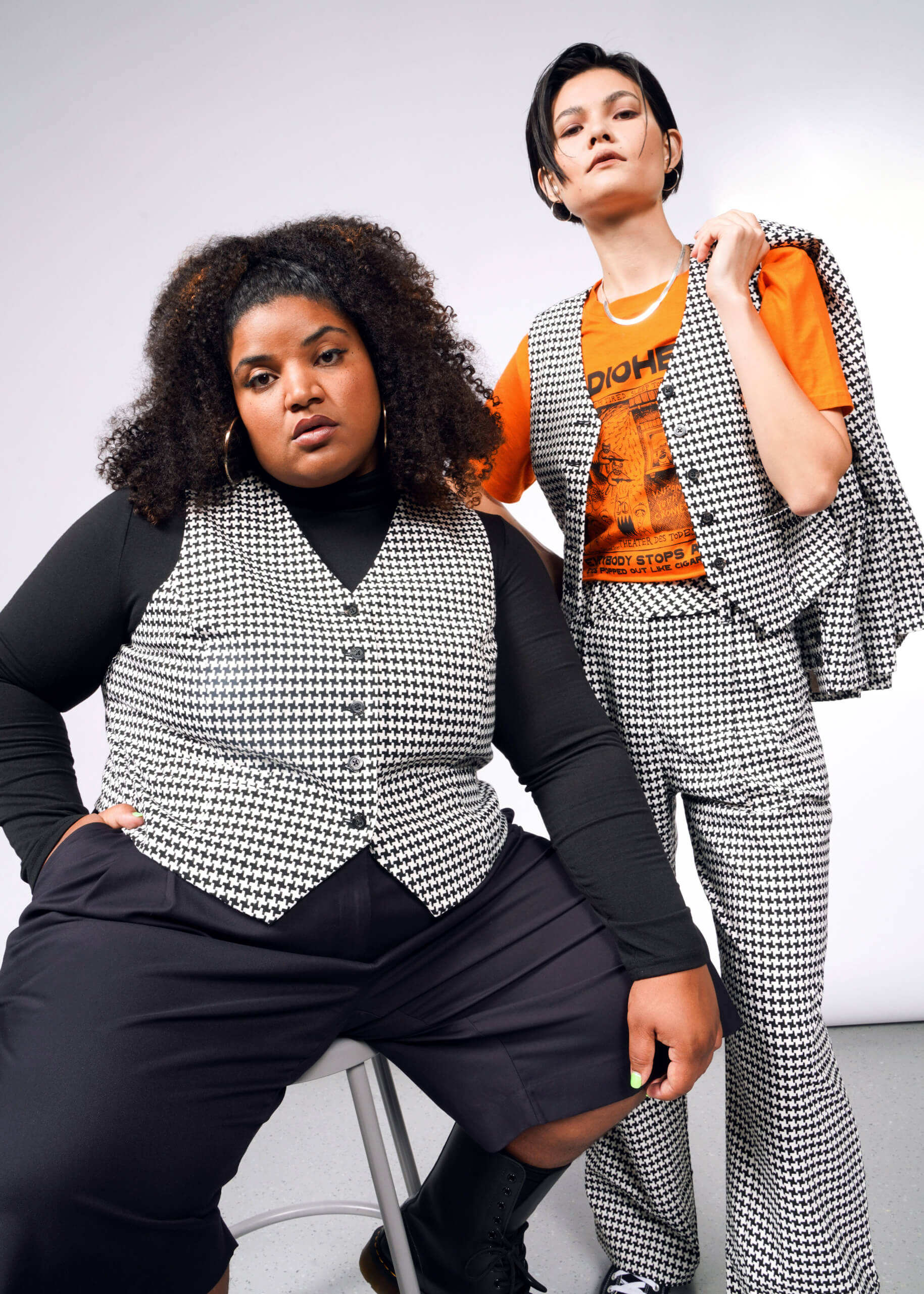 Two people wear matching houndstooth outfits. One has a tailored look with The Empower Houndstooth Vest over an orange graphic tee and flared pants; the other sits on a stool in boots. A neutral gray background enhances their coordinated style.