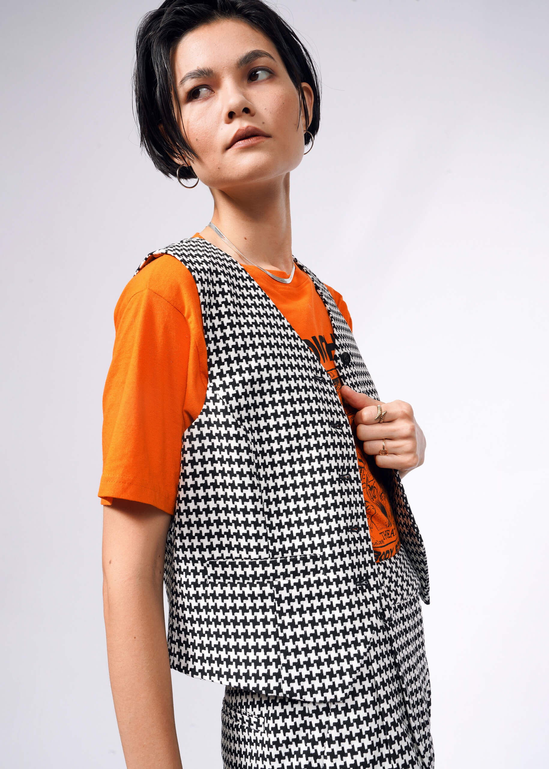 Sporting short black hair, the individual wears The Empower Houndstooth Vest—perfect for layering due to its tailored fit—with matching pants over an orange t-shirt, gazing sideways.