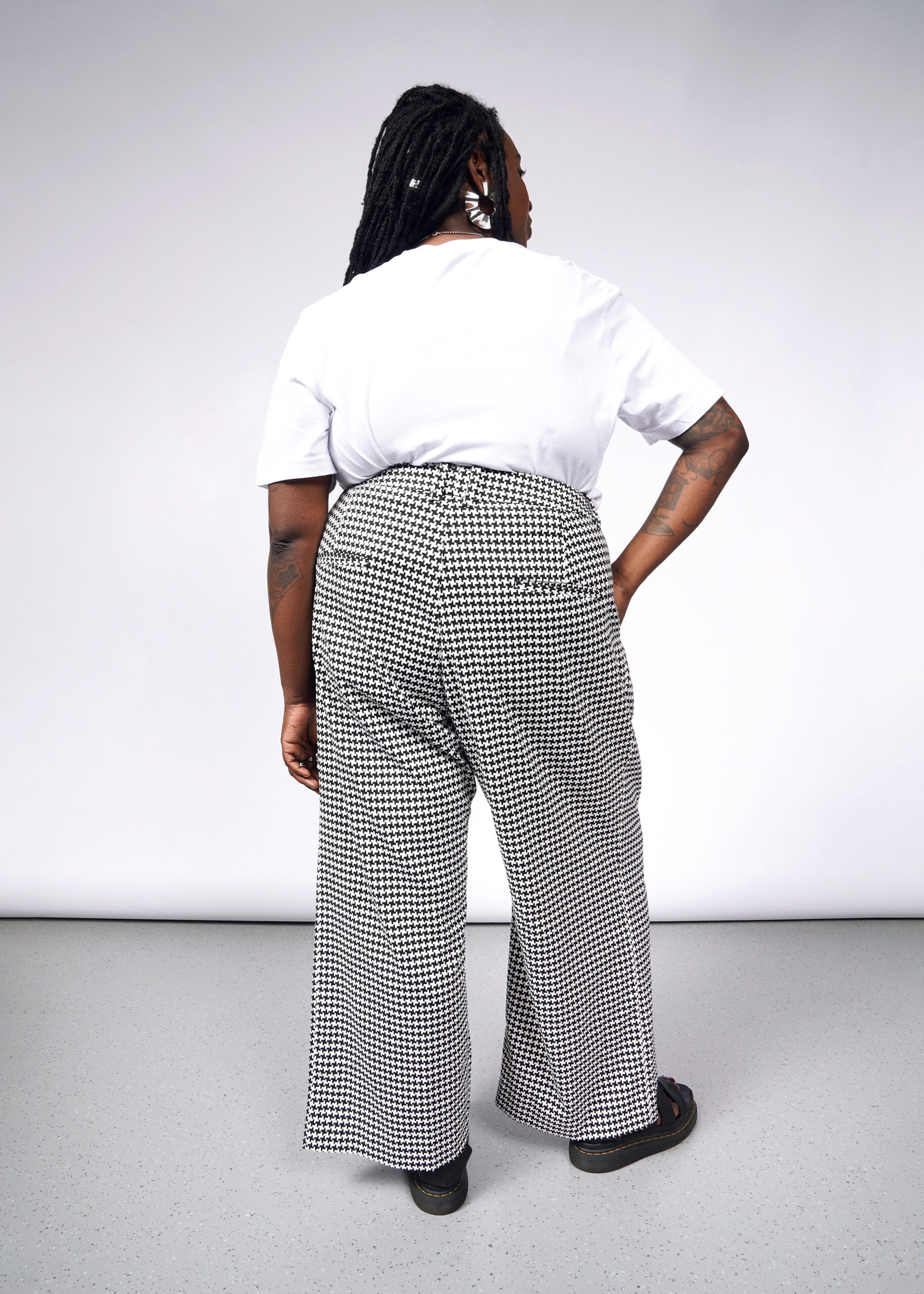 With long braided hair and arms adorned with tattoos, a person stands back to the camera against a light gray background, showcasing The Empower Houndstooth Wide Leg Trouser, styled with a white t-shirt and black sandals.