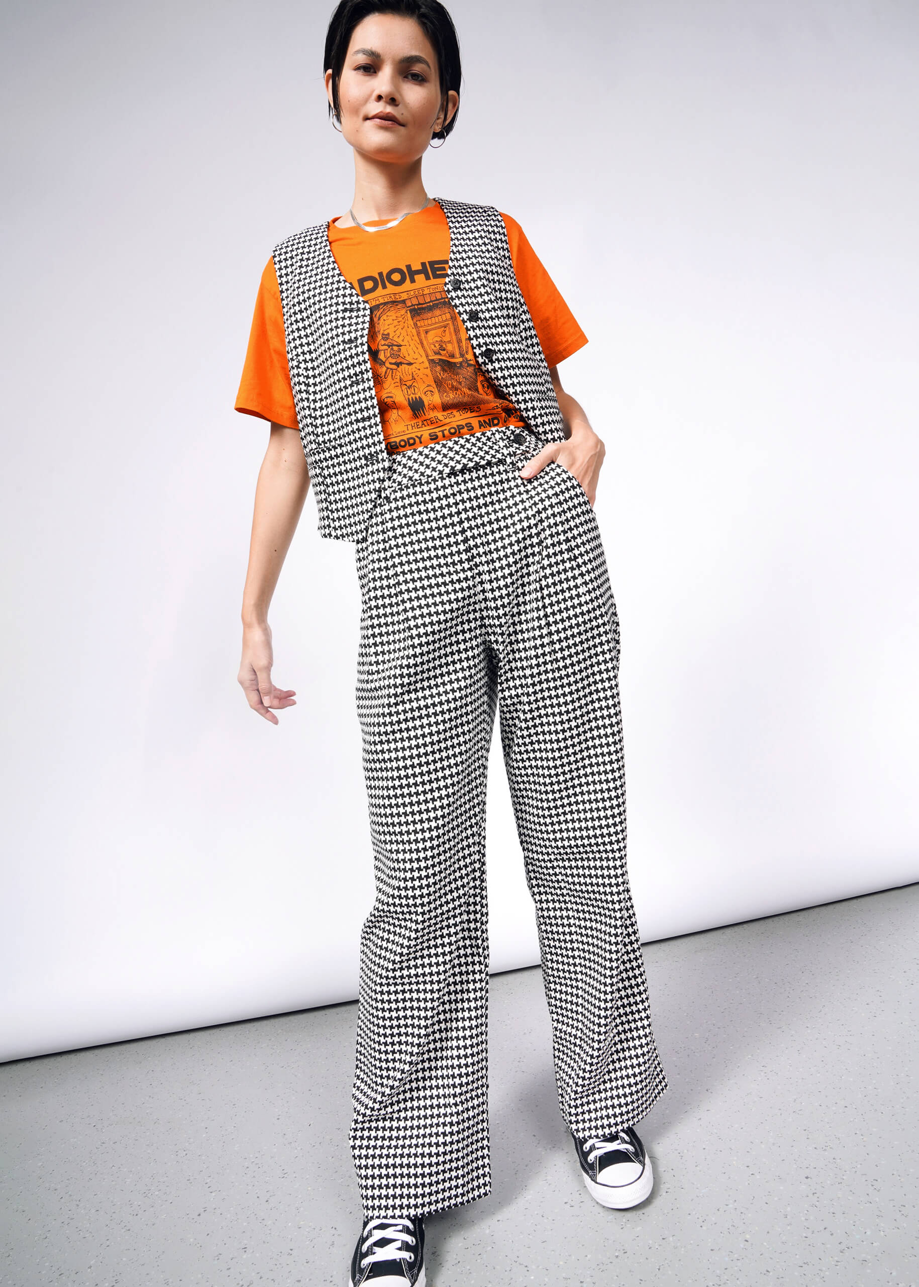 A person exudes confidence in high-waisted trousers, showcasing The Empower Houndstooth Wide Leg Trouser and a matching vest, paired with an orange graphic t-shirt. Black and white sneakers complete the ensemble against a plain gray backdrop, highlighting their chic style.