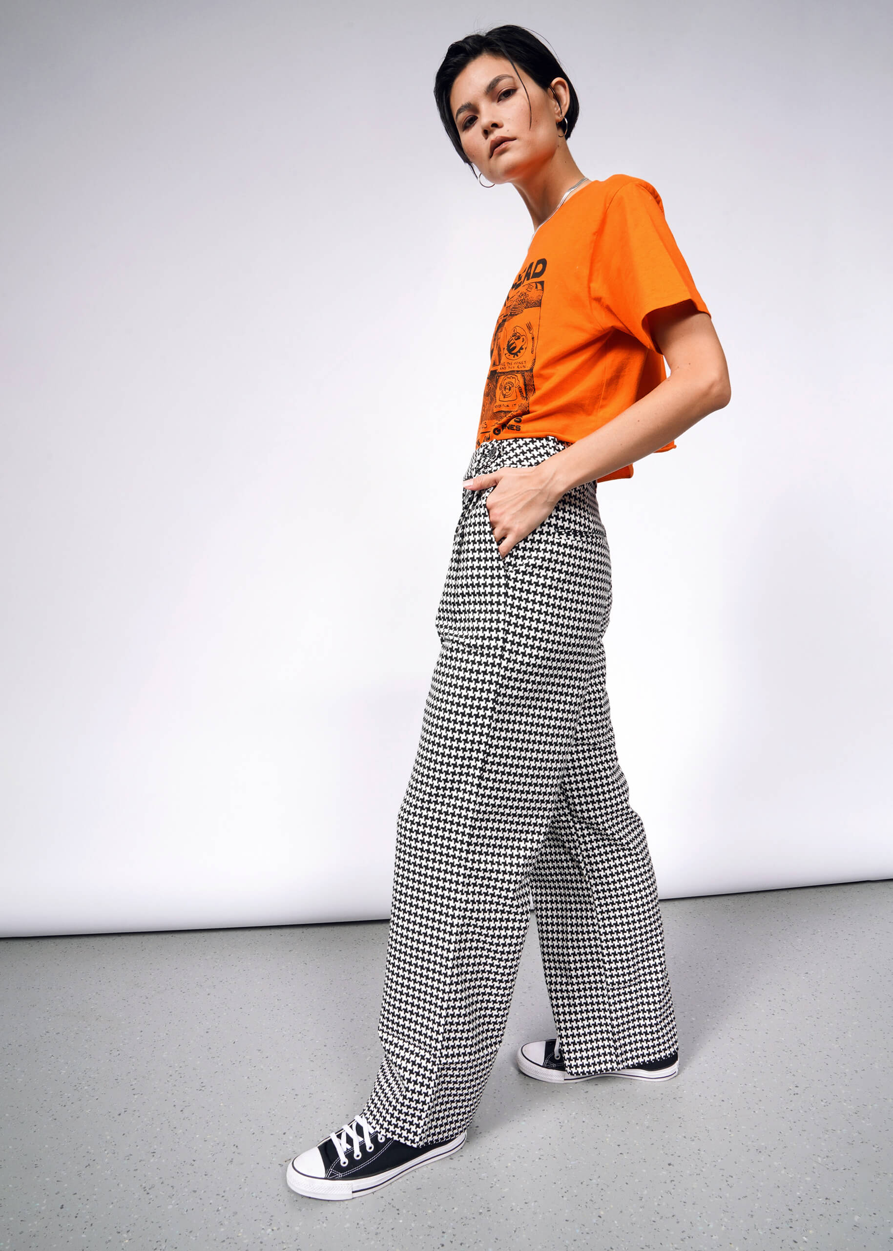 A person with short dark hair stands against a plain background, wearing an orange graphic T-shirt paired with The Empower Houndstooth Wide Leg Trouser in black and white checkered with double front pleats. They have their hands in pockets, black sneakers on, and are looking to the side.