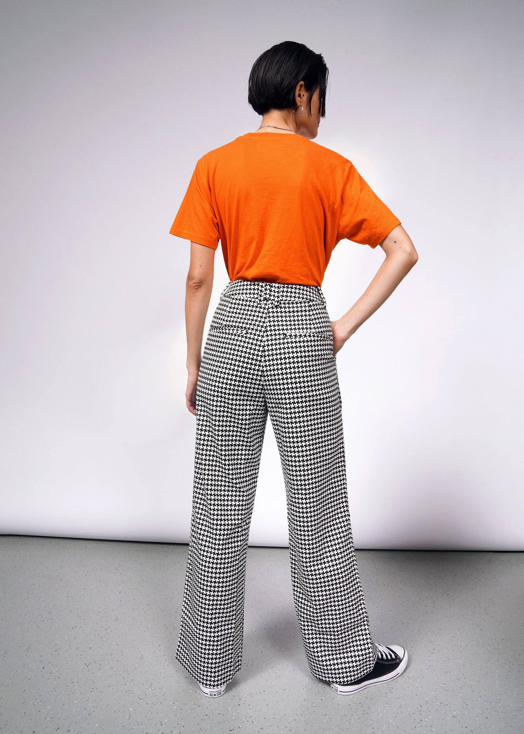 A person with short dark hair stands facing away in a minimalist setting with a neutral background, wearing an orange T-shirt and The Empower Houndstooth Wide Leg Trouser. One hand rests on their hip.