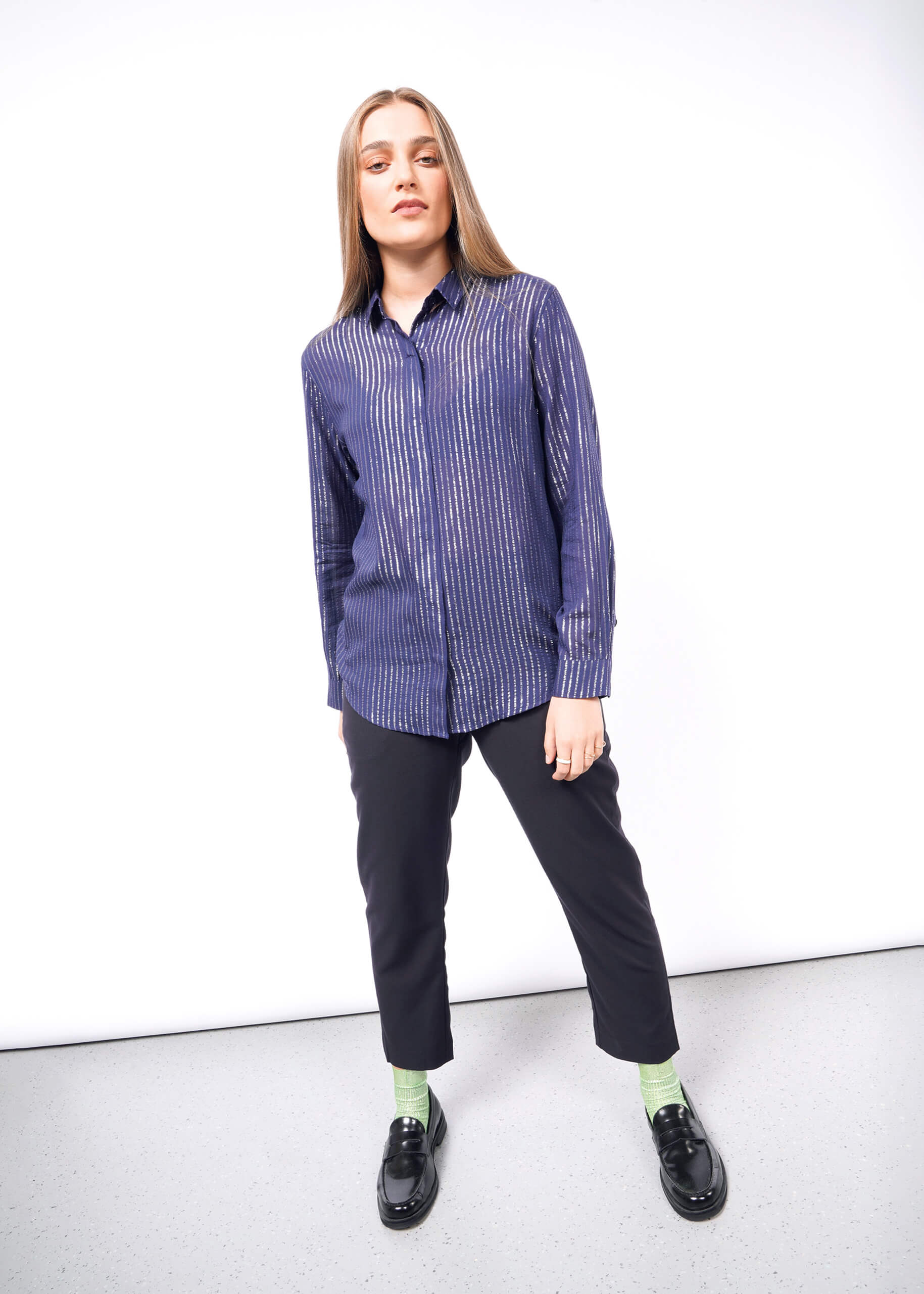 A person with long hair stands relaxed against a white backdrop, wearing The Empower Sheer Pinstripe Long Sleeve Button Up, black pants, green socks, and black shoes.