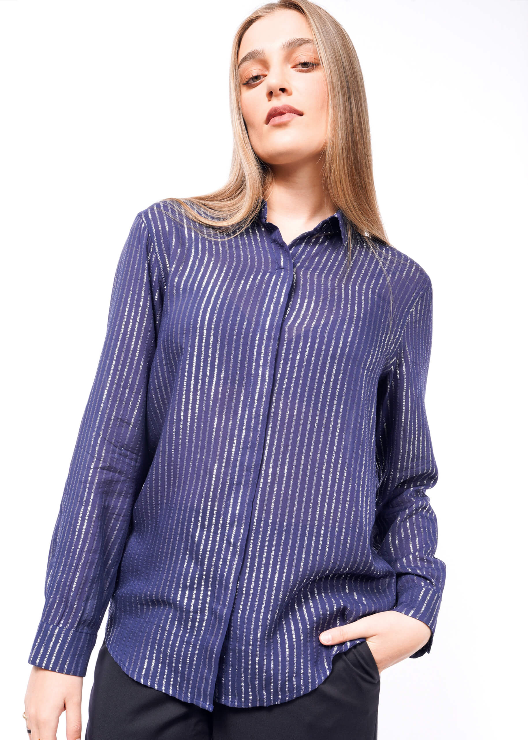 A person with long hair wearing The Empower Sheer Pinstripe Long Sleeve Button Up, characterized by a relaxed fit and metallic pinstripes, tilts her head and poses with one hand in a pocket against a plain white backdrop.