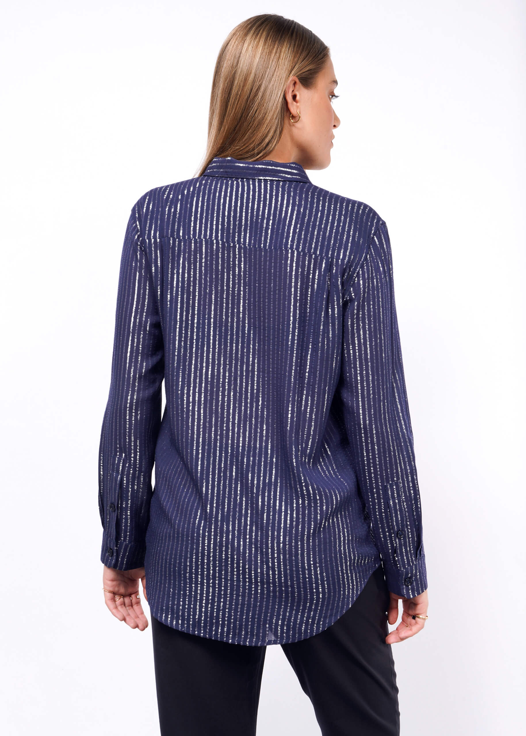 A person with long hair, shown from behind, wears The Empower Sheer Pinstripe Long Sleeve Button Up in dark blue and metallic pinstripes paired with black pants; the relaxed fit complements the plain white background.