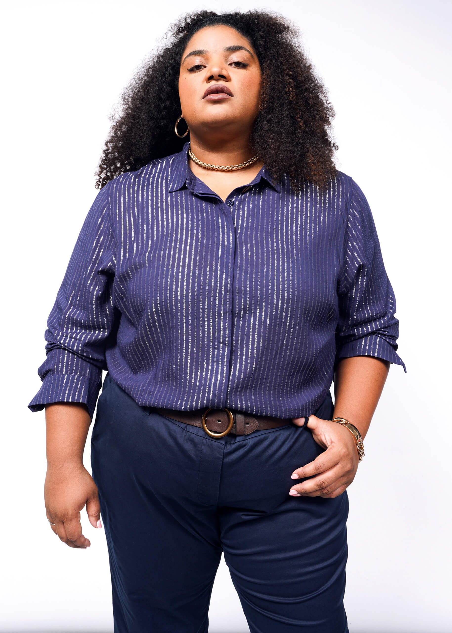 Person with long curly hair wearing The Empower Sheer Pinstripe Long Sleeve Button Up in blue and dark pants, standing confidently against a white background. They have a gold chain necklace and a brown belt.