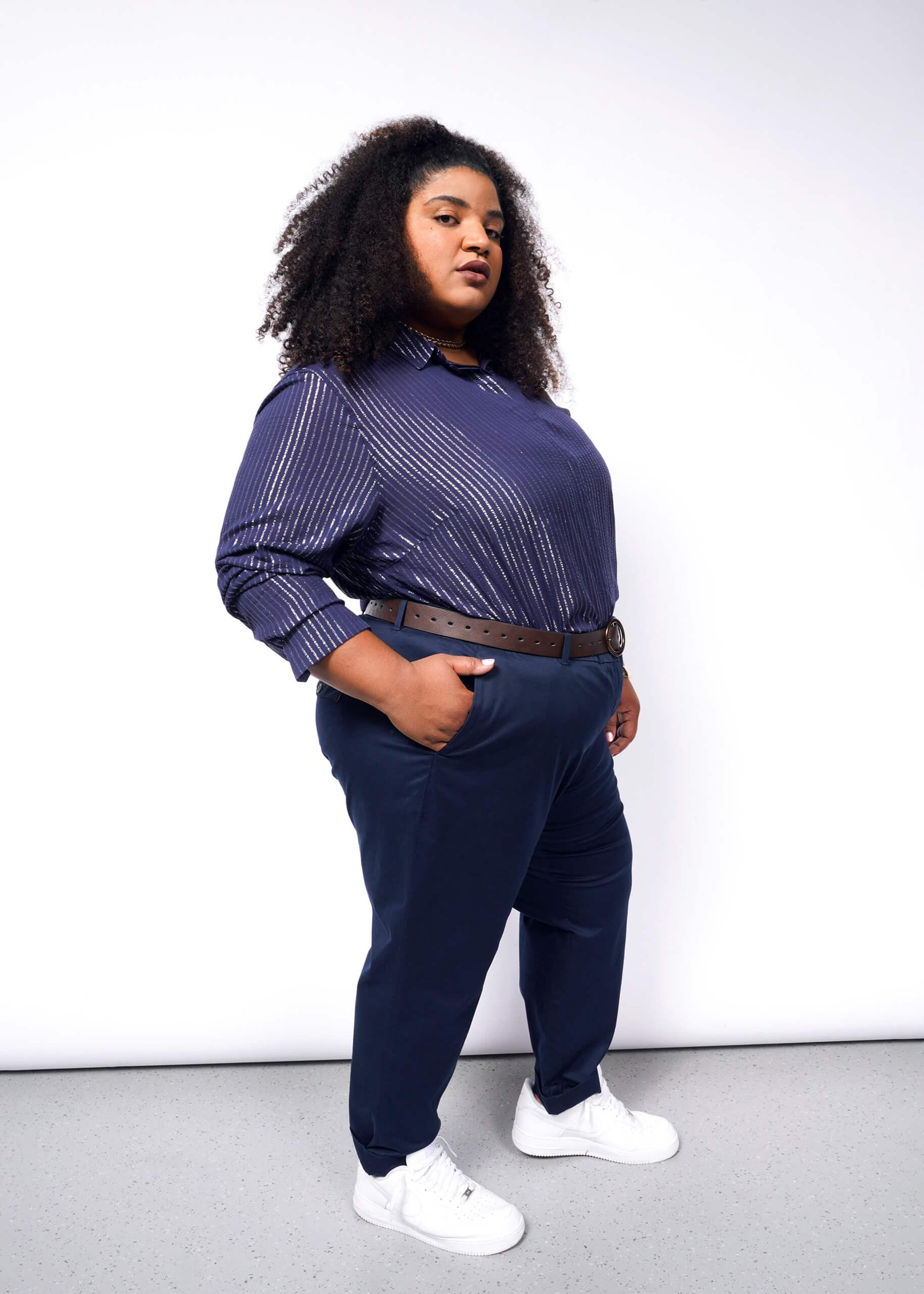 Wearing The Empower Sheer Pinstripe Long Sleeve Button Up and relaxed fit dark blue pants, a person with long, curly hair stands confidently against a white background. Their hands are in their pockets, and they have a belt with a round buckle complementing their white sneakers.