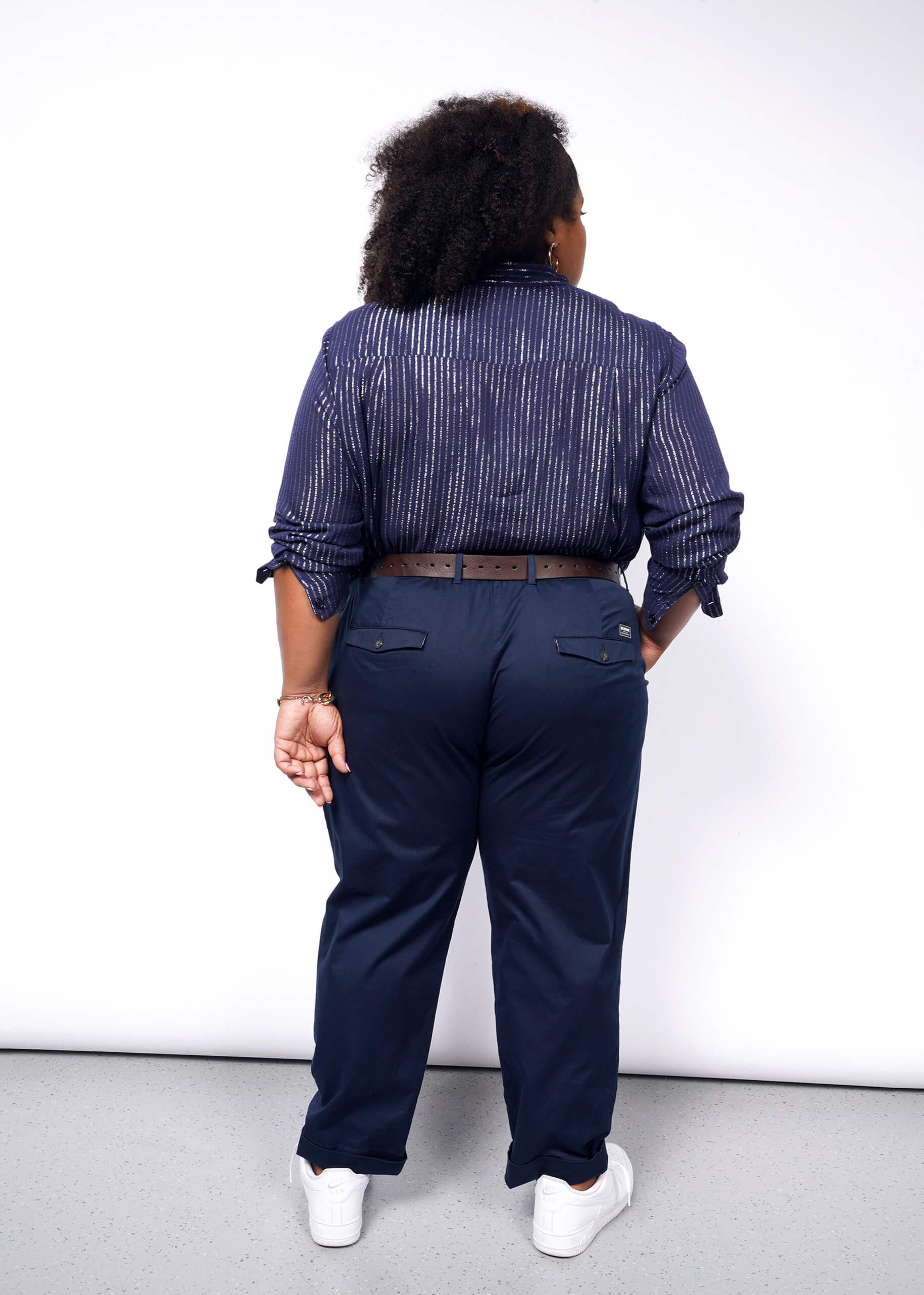 A person with curly hair stands with their back to the camera, wearing The Empower Sheer Pinstripe Long Sleeve Button Up and relaxed-fit navy pants, complemented by a brown belt. They sport white sneakers in a minimalist white background setting.