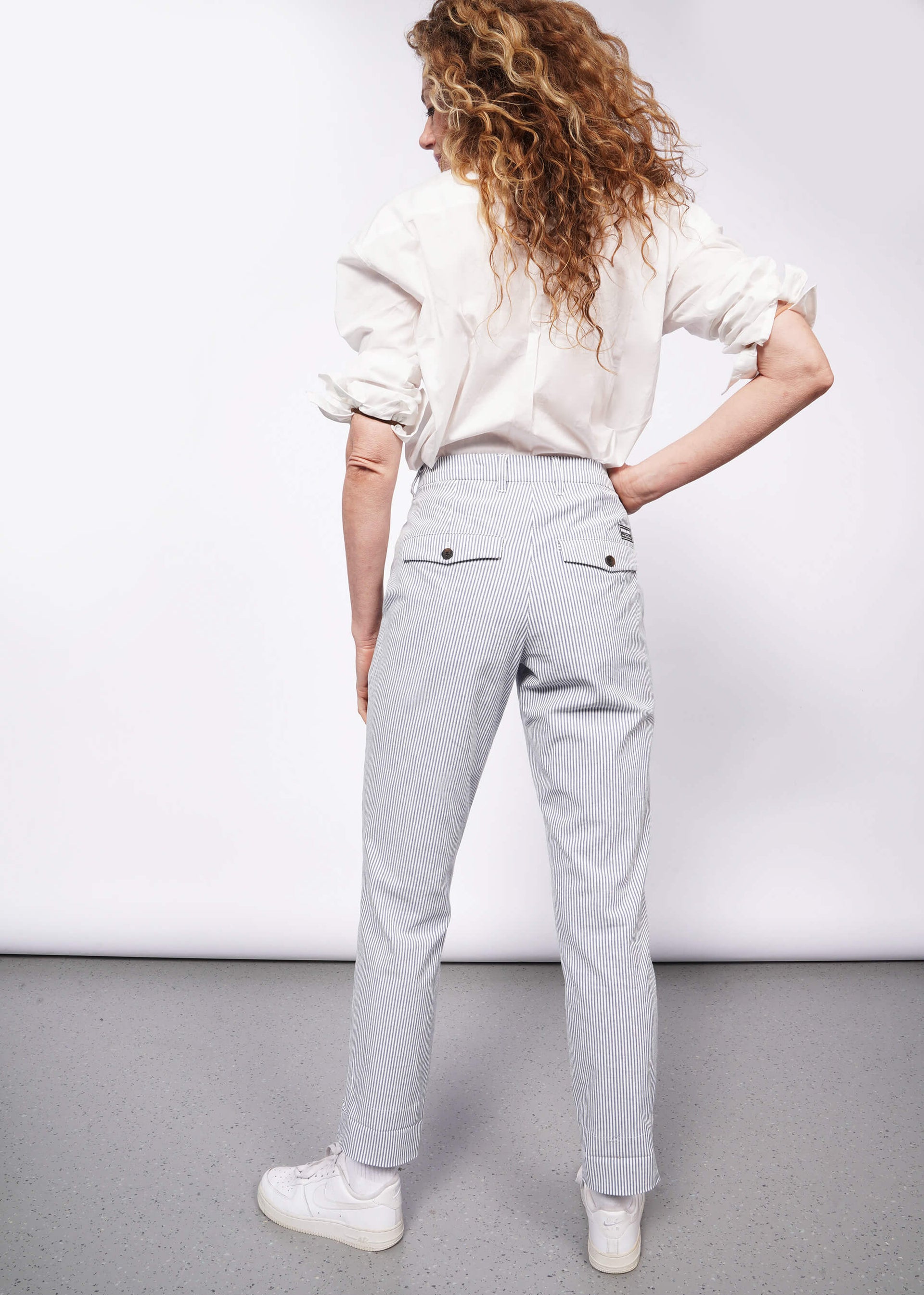 Back of person with wavy blonde hair with hand on hip wearing white long sleeve button up tucked into seersucker white and blue trousers with white sneakers.