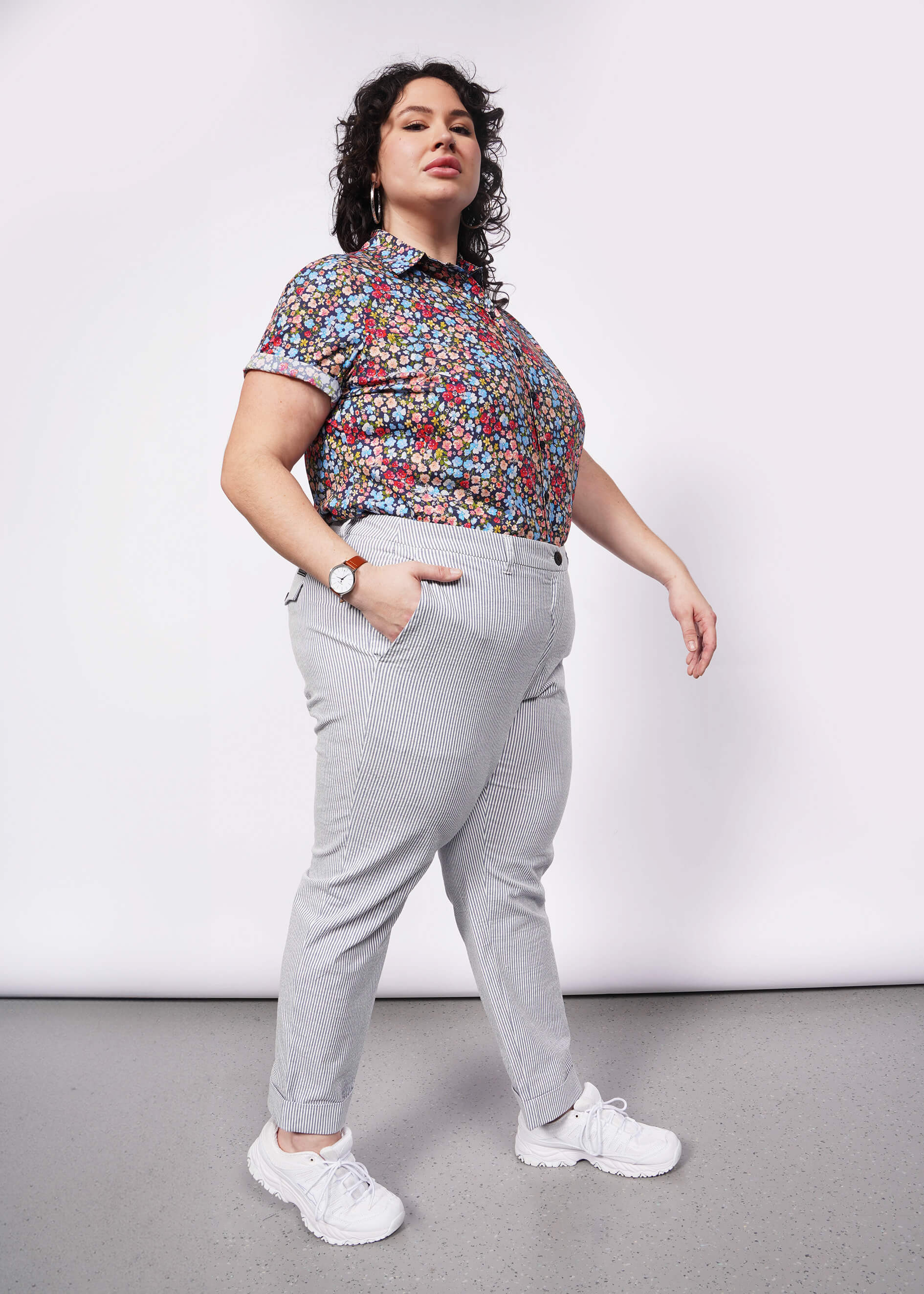 Side view of model with curly black hair wearing floral printed button up tucked into seersucker blue and white trouser with white sneakers.