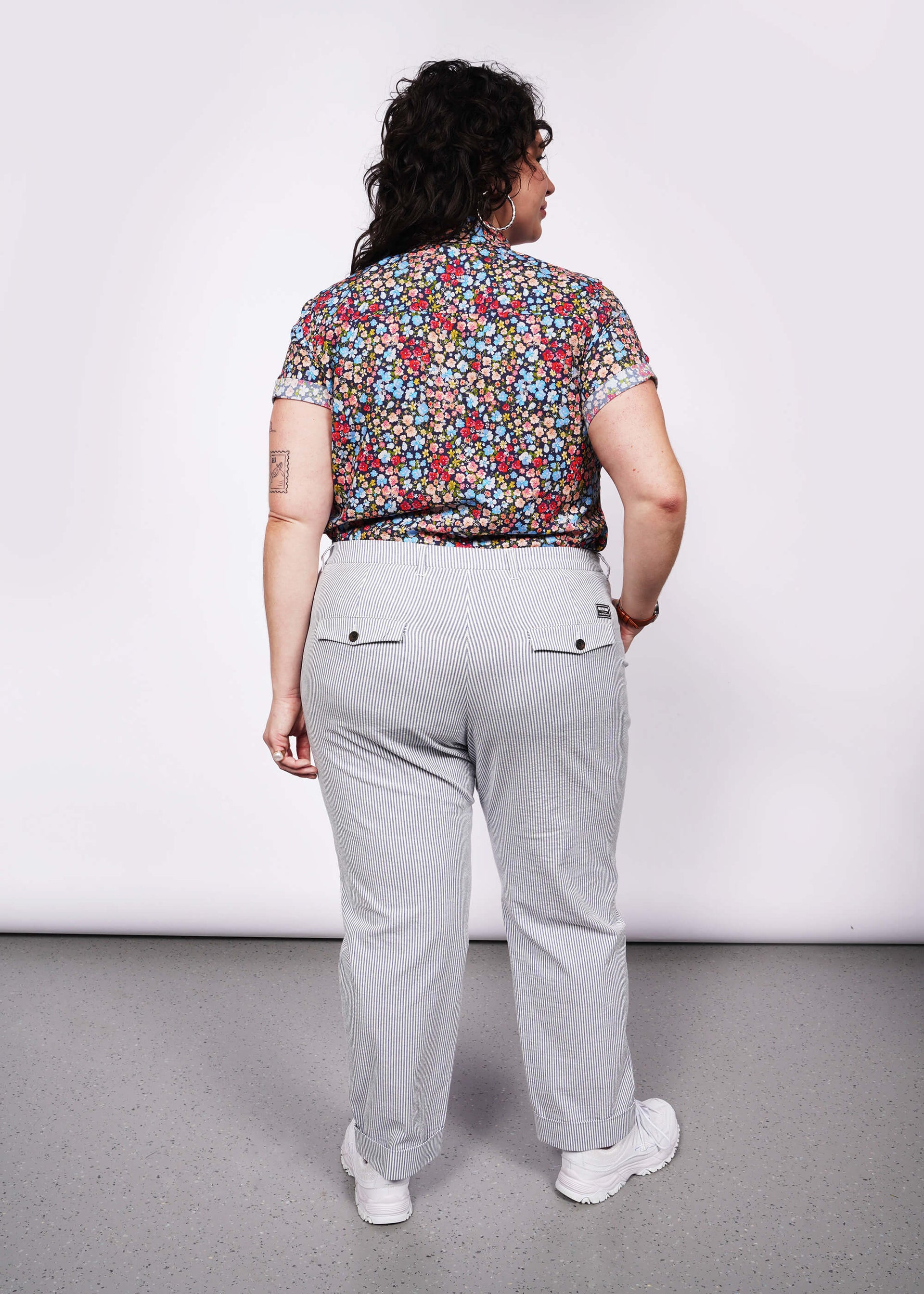 Back view of model with curly black hair wearing floral printed button up tucked into seersucker blue and white trouser with white sneakers.