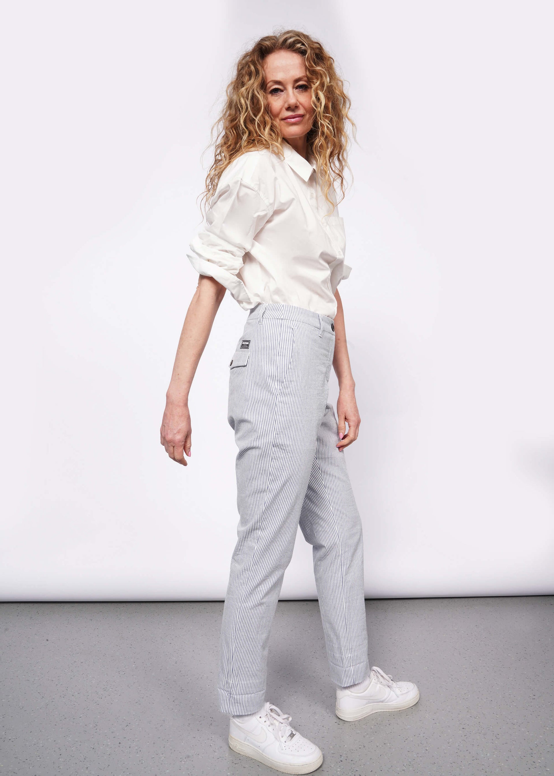 Person with wavy blonde hair stands to the side whole looking at camera wearing white long sleeve button up tucked into seersucker white and blue trousers with white sneakers.